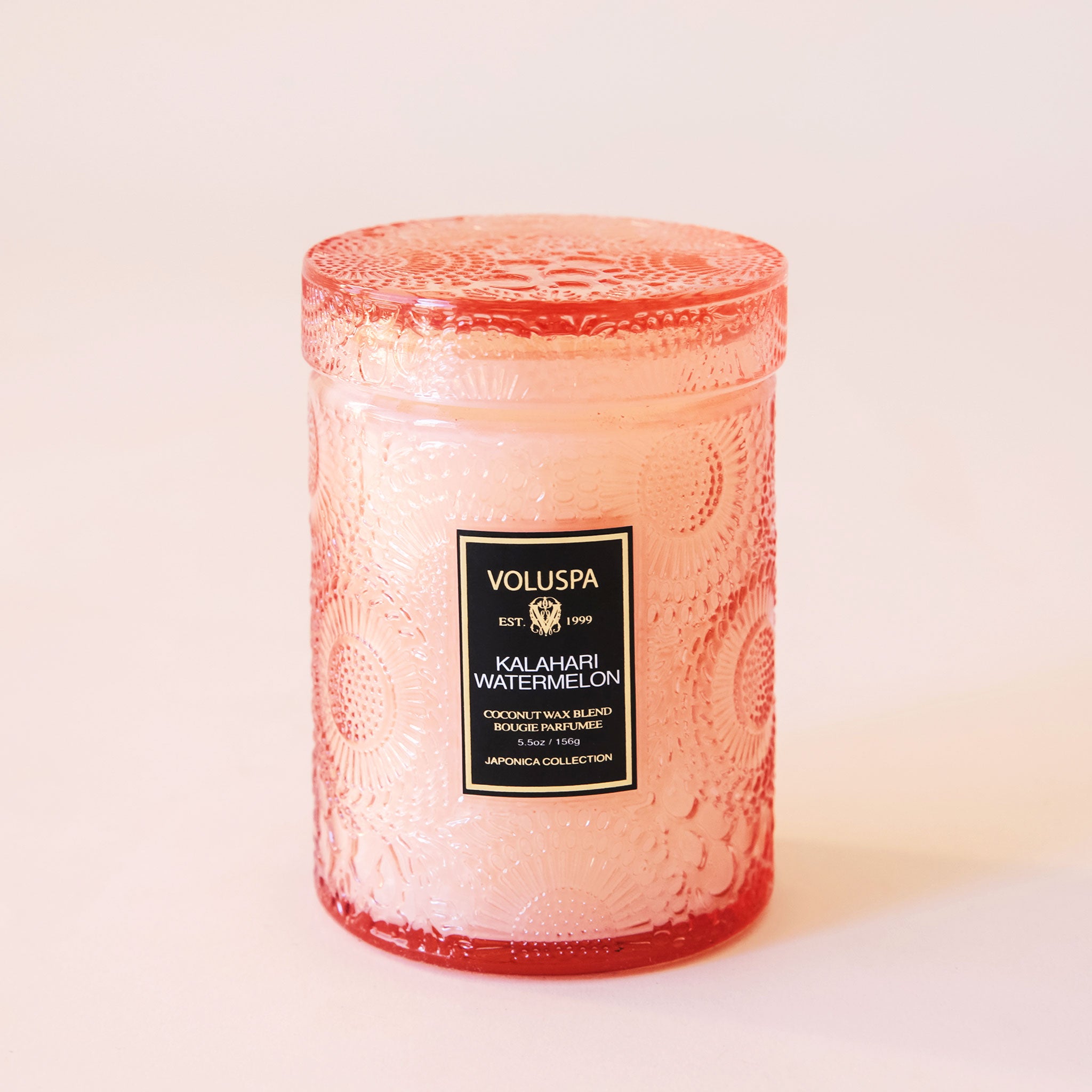 A peach colored embossed jar with an abstract floral and spotted design. The glass is somewhat clear, but is thick in quality making it slightly opaque. Its lid is also glass and has the same embossing design. The voluspa label is black with gold lettering.