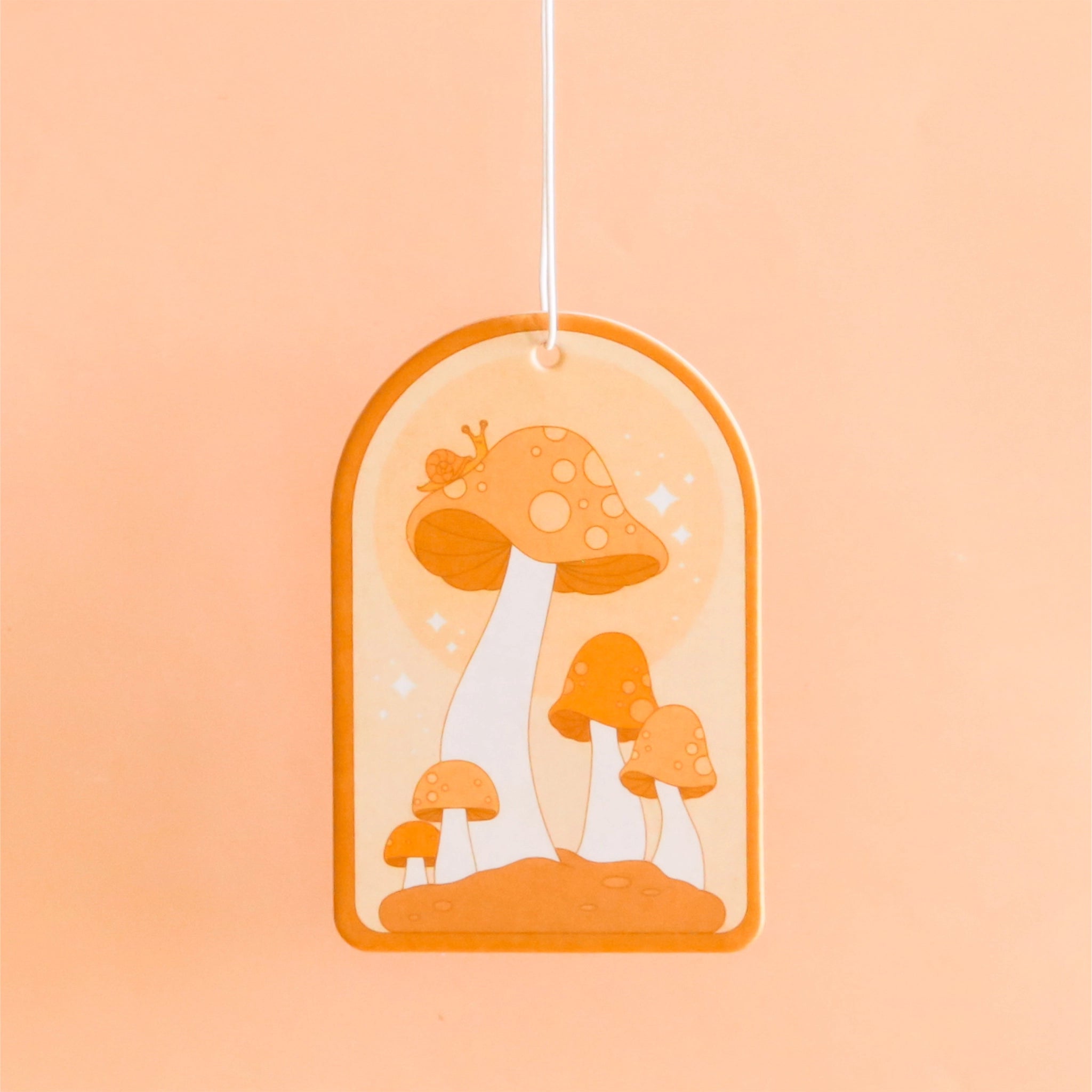 On a light orange background is an arched shaped air freshener with an orange mushroom design and a white elastic loop for hanging.