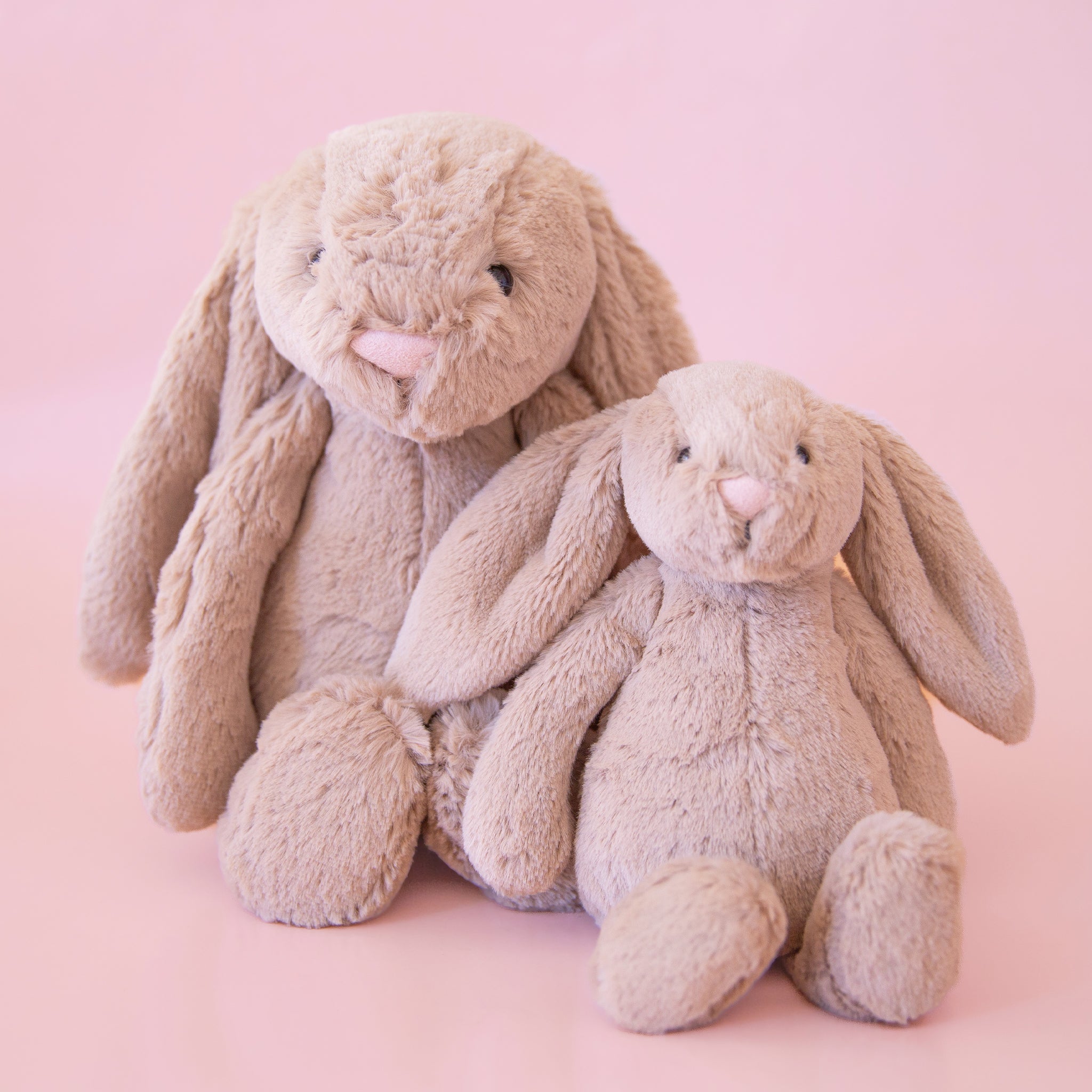 Two, one small and one large, stuffed animals in the shape of beige-grey colored rabbits, with long floppy ears, arms and legs.