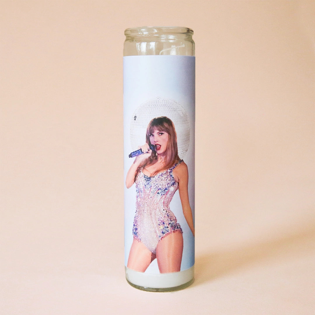 On a tan background is a prayer candle featuring a light blue background and the iconic Taylor Swift on the front in a sparkly onesie. 