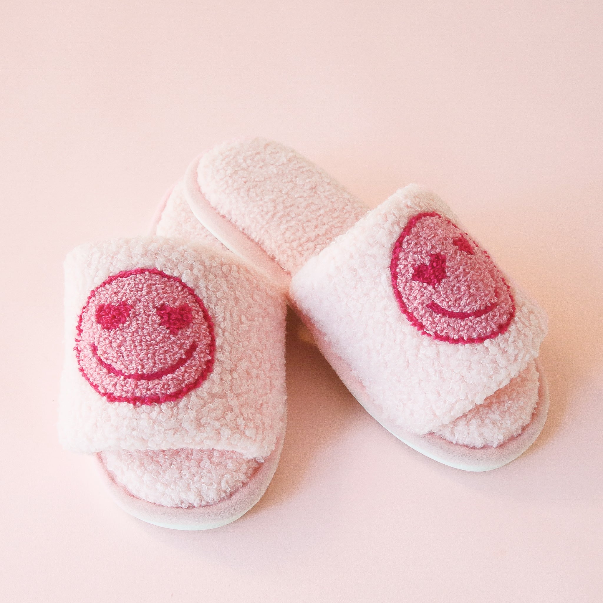 Pink fuzzy slide slippers with a darker pink heart eyes smiley face graphic on the front of each slipper.
