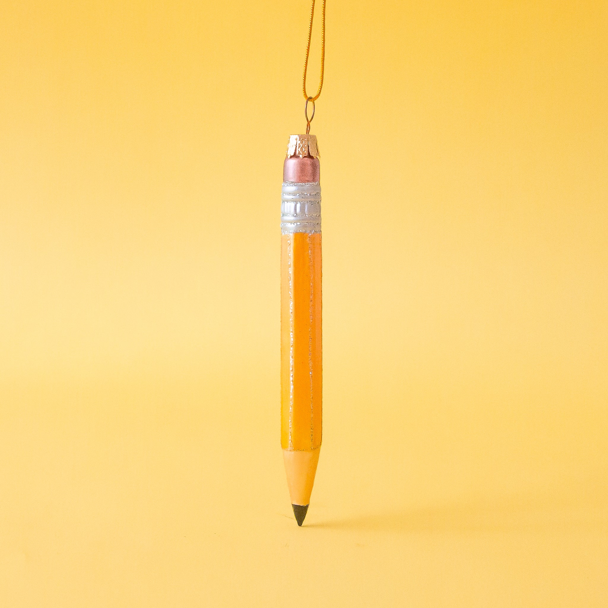 On a yellow ground is a photo of a pencil shaped Christmas ornament. The pencil has a pink eraser, black tip and a gold string hanging at top to hang on a tree.