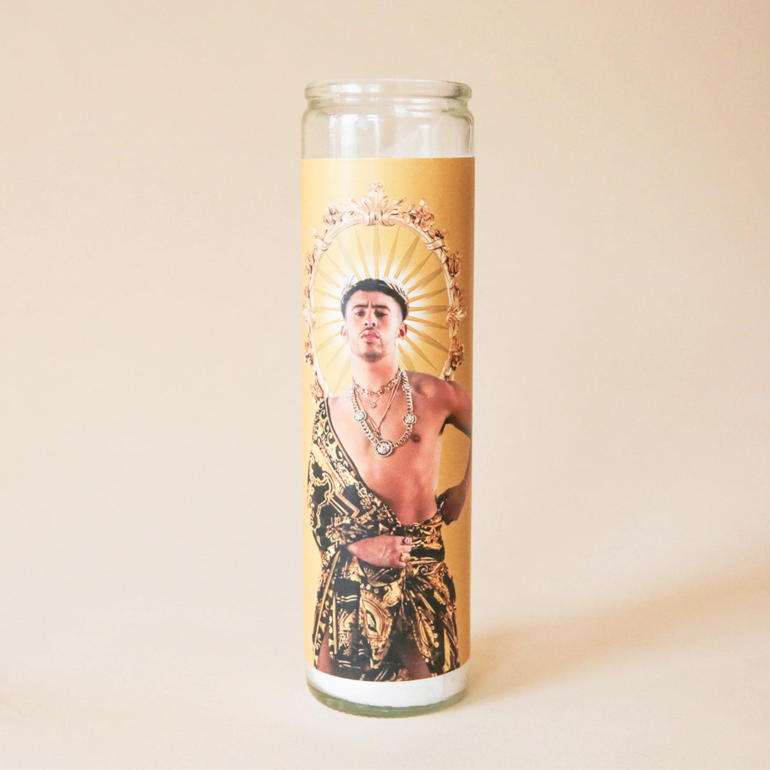 On a tan background is a thin, gold prayer candle with a photo of Bad Bunny. 