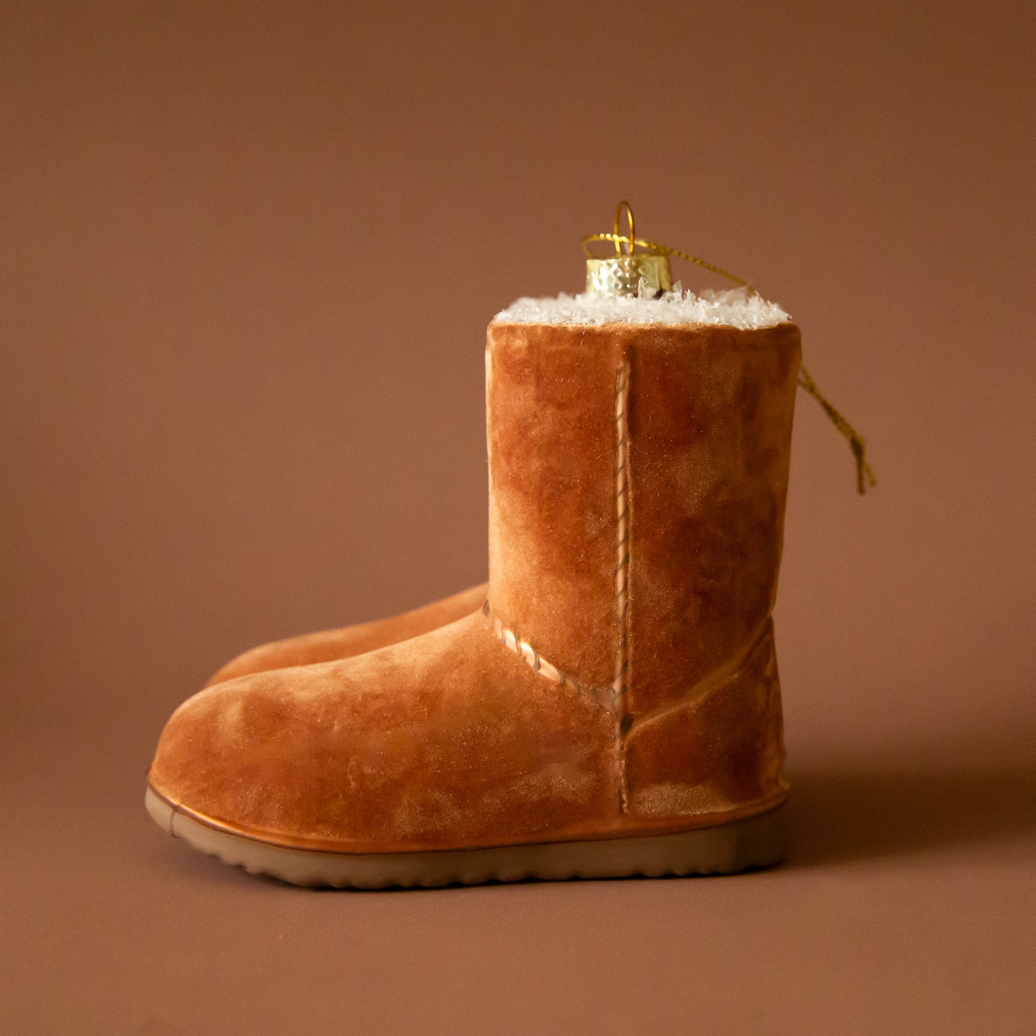a pair of tan velvet UGG boots on a brass ring on a brown background. The boots are a Christmas ornament and have brown stitching detail.