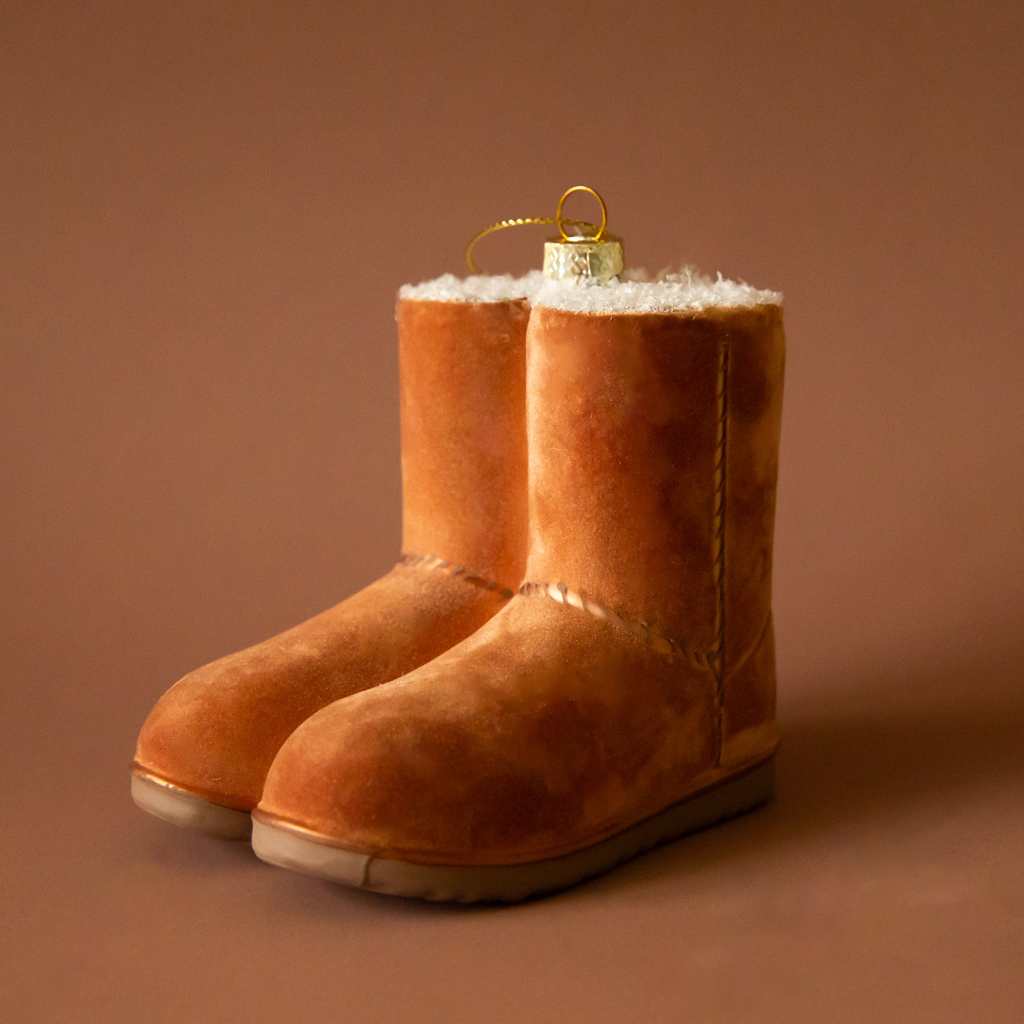 A pair of tan velvet UGG boots on a brass ring on a brown background. The boots are a Christmas ornament and have brown stitching detail.