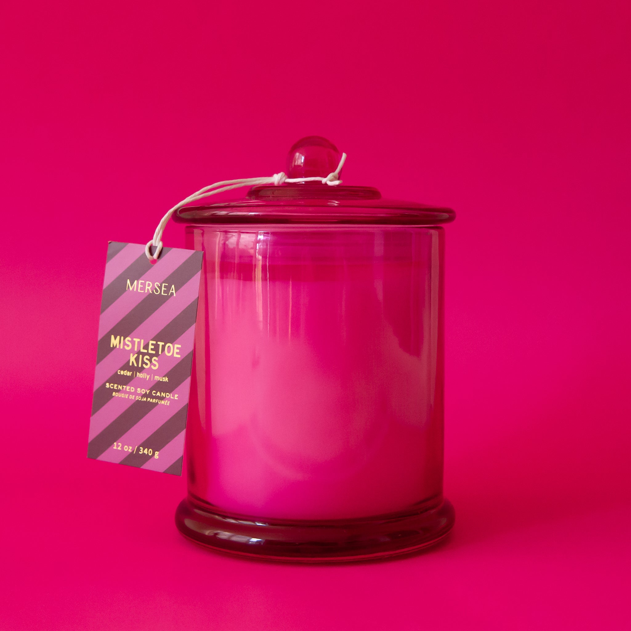 A beautiful hot pink glass cylinder shaped candle sits in the middle of image.  The lid has a little round knob with a paper tag hanging off that reads "Mistletoe Kiss".  The label has purple and pink stripes and hangs from a white string tied around the round knob.  The background of the image is also hot pink.