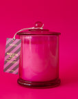 A beautiful hot pink glass cylinder shaped candle sits in the middle of image.  The lid has a little round knob with a paper tag hanging off that reads "Mistletoe Kiss".  The label has purple and pink stripes and hangs from a white string tied around the round knob.  The background of the image is also hot pink.