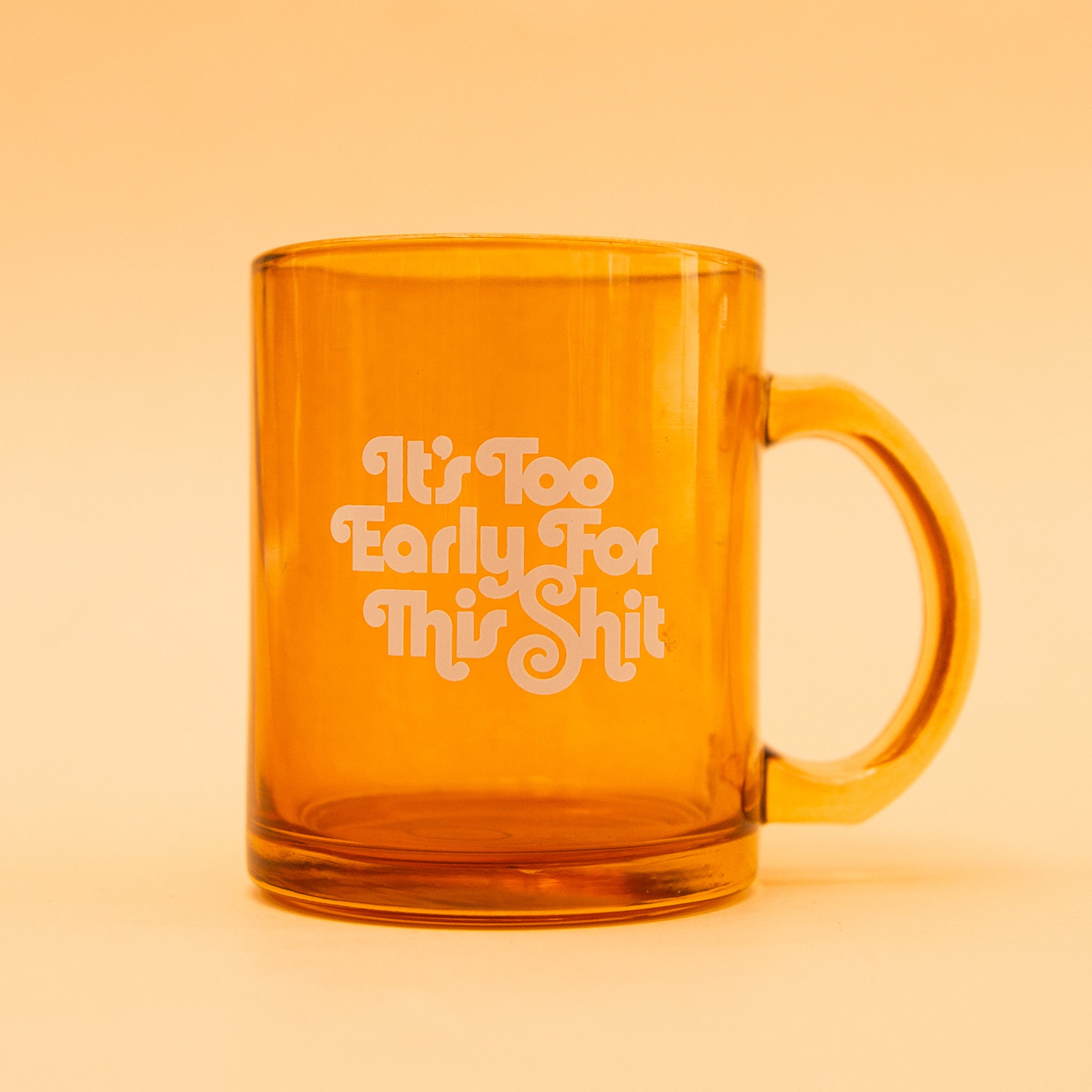 On a yellow background is an orange glass mug with text that reads, "It's Too Early For This Shit". 