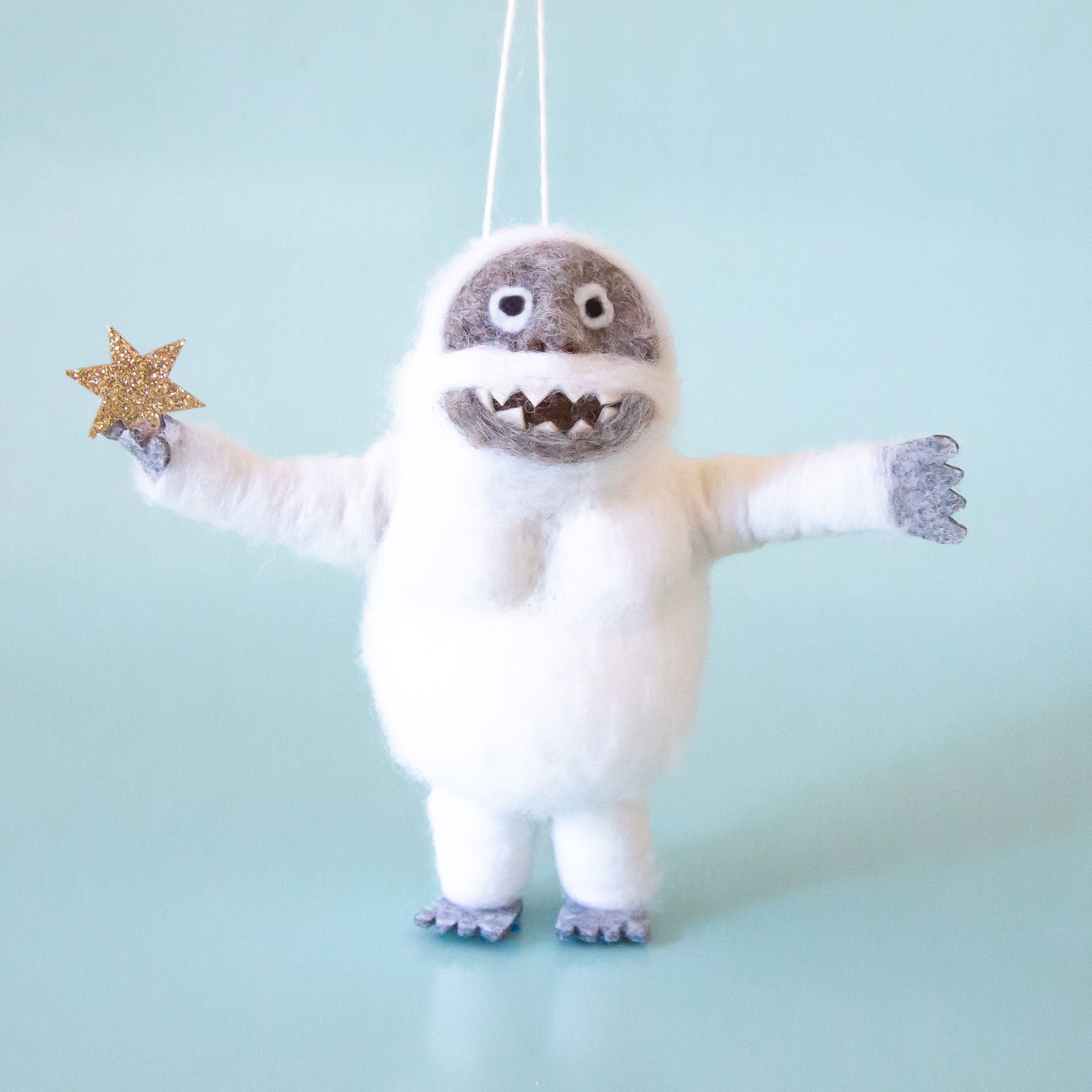 On a light blue background is a white and grey felt abominable snowman ornament that's holding a sparkly star