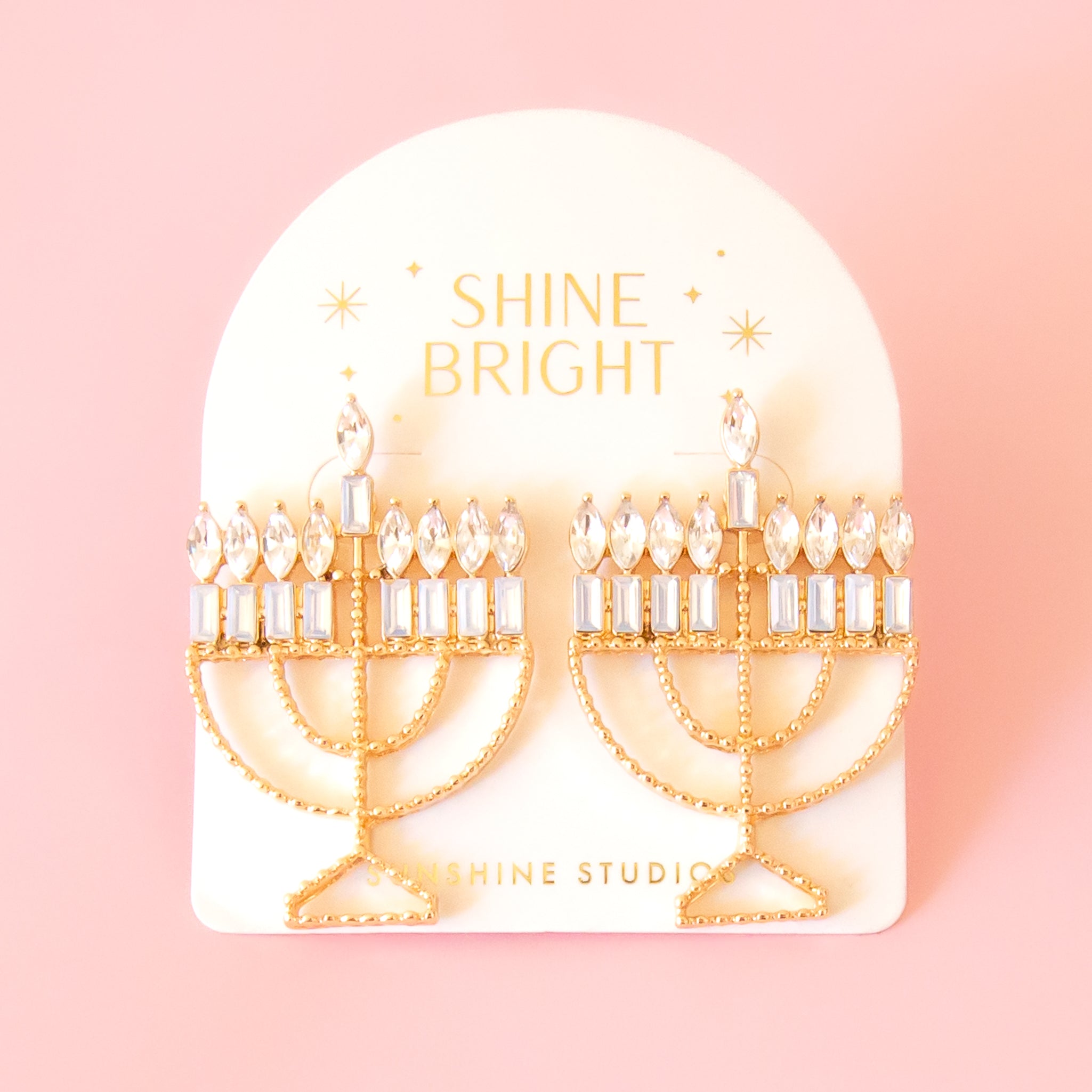Menorah shaped gold earrings are attached to a cream colored card. the Card has gold foil stamp which reads Shine Bright and Sunshine Studios with glittering stars surrounding the text. The background of the photo is a soft warm pink color. 