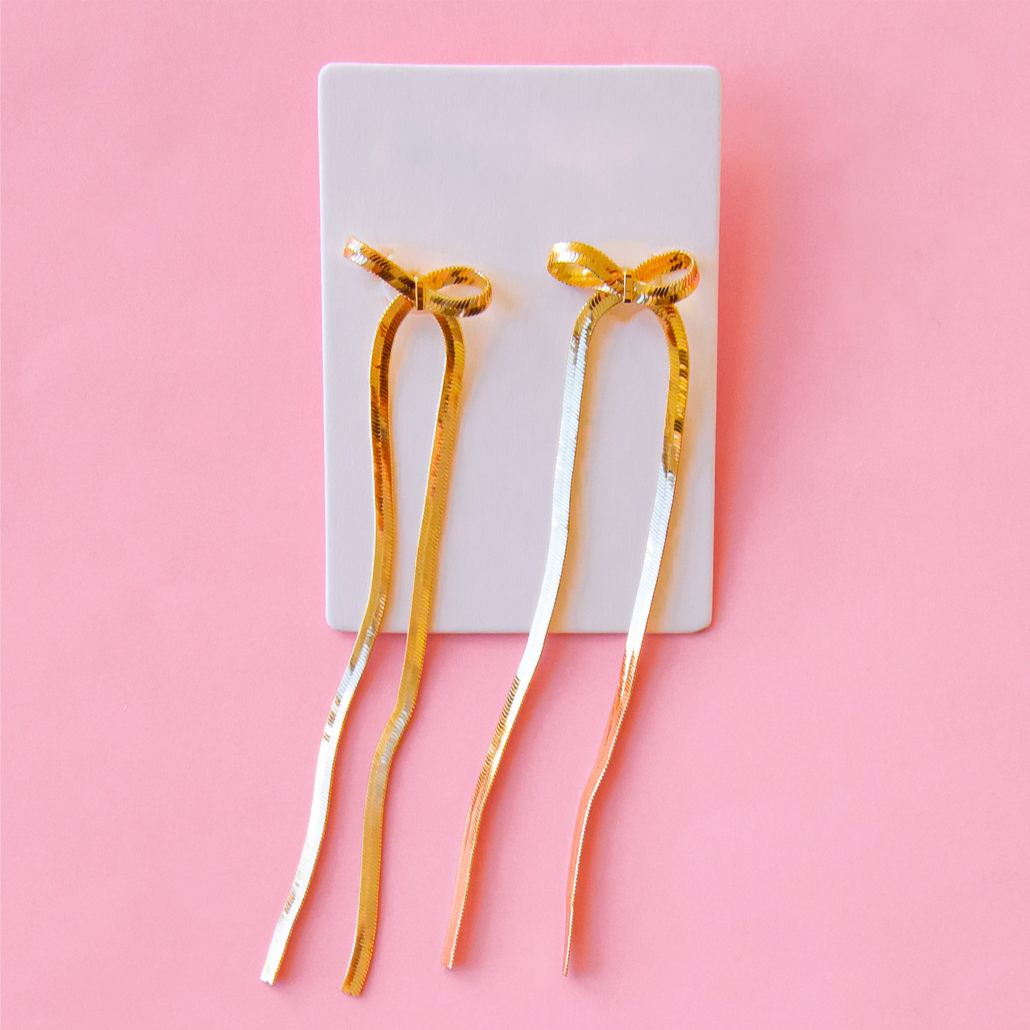On a pink background is a pair of gold dangle bow shaped earrings. 