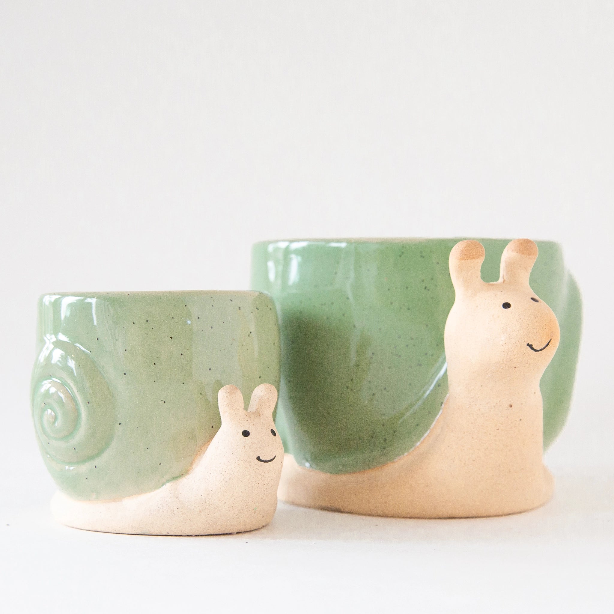 On a light grey background is two ceramic planters in the shape of a snail with smiling faces and a swirly green &quot;shell&quot;.
