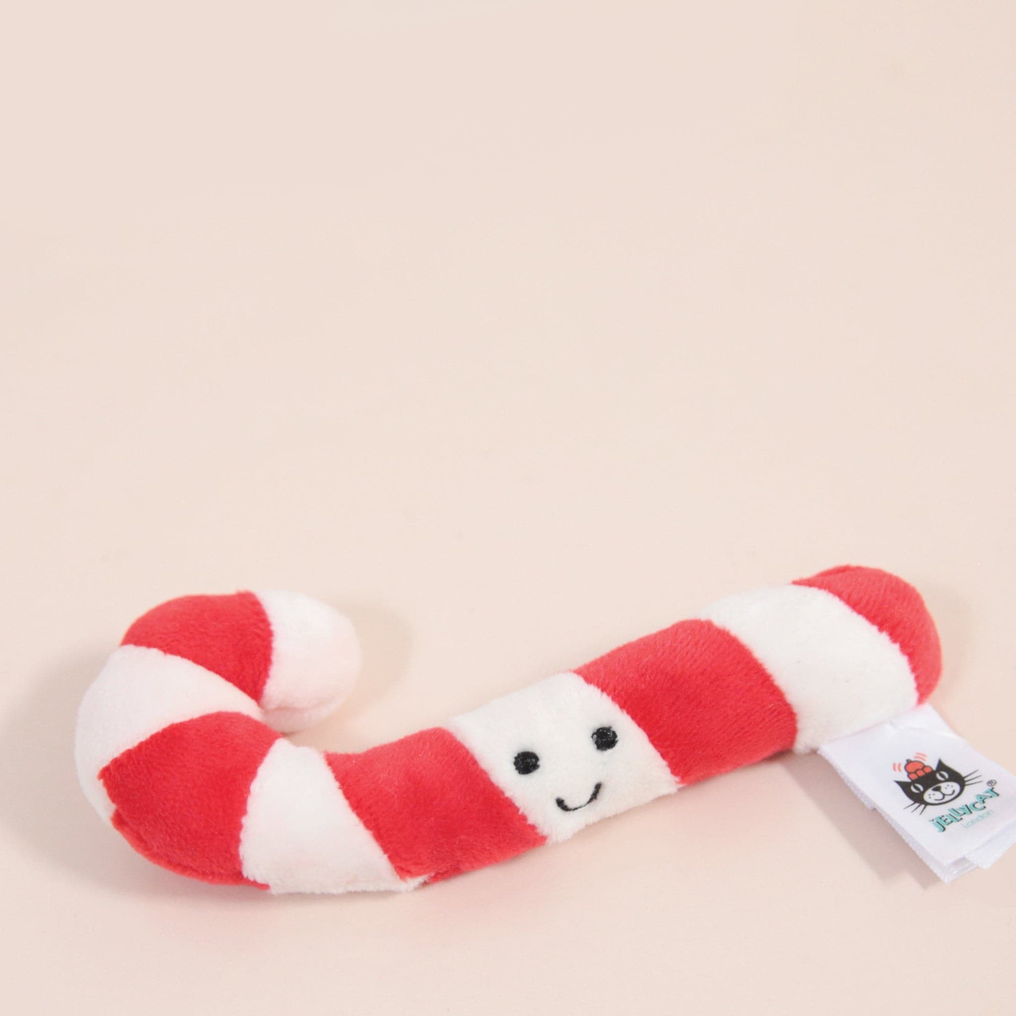 On a light pink background is a red and white candy cane stuffed toy with a smiling face in the center. 