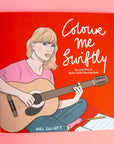 On a pink background is a red book with an illustration of Taylor Swift with a guitar and the white title on the right that reads, "Colour me Swiftly The Unofficial Taylor Swift Colouring Book".