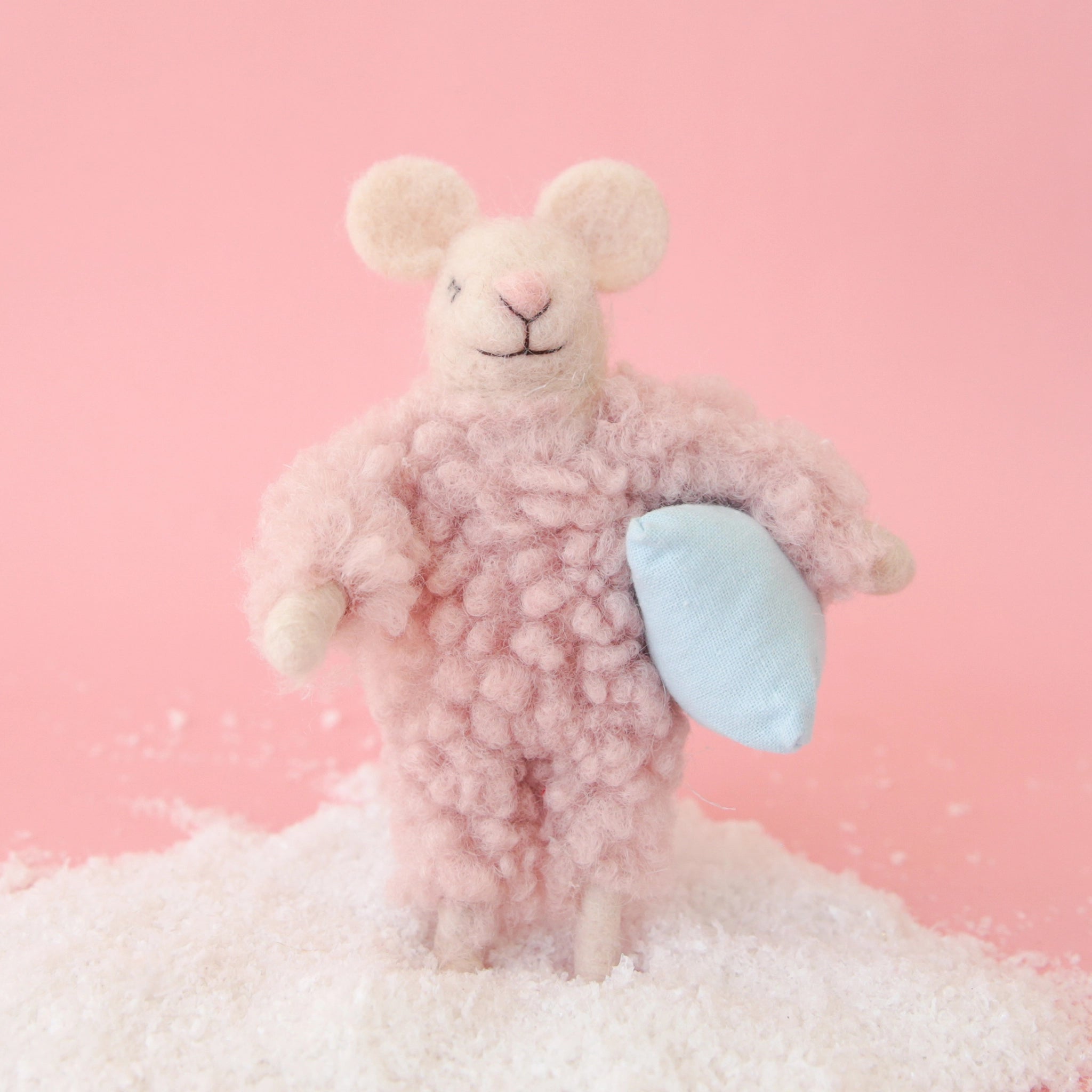On a pink background is a felt mouse ornament in a cream shade with a fuzzy pink jumpsuit on and holding a pillow.