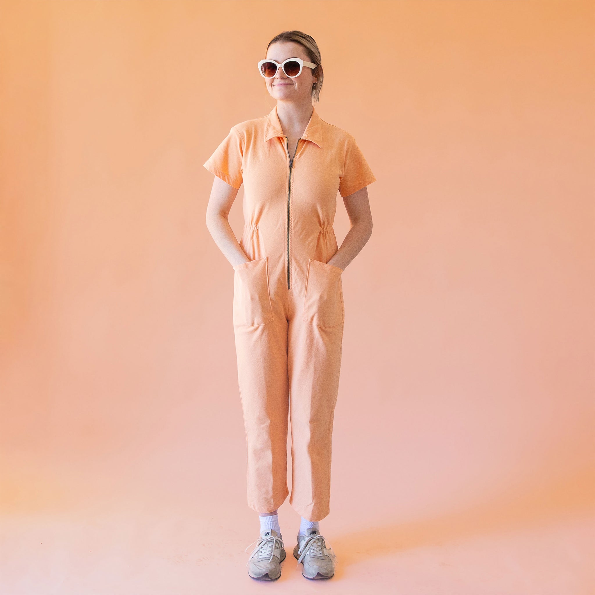 A model wearing a light peach colored short sleeve jumpsuit with a zipper up the front and a collar.