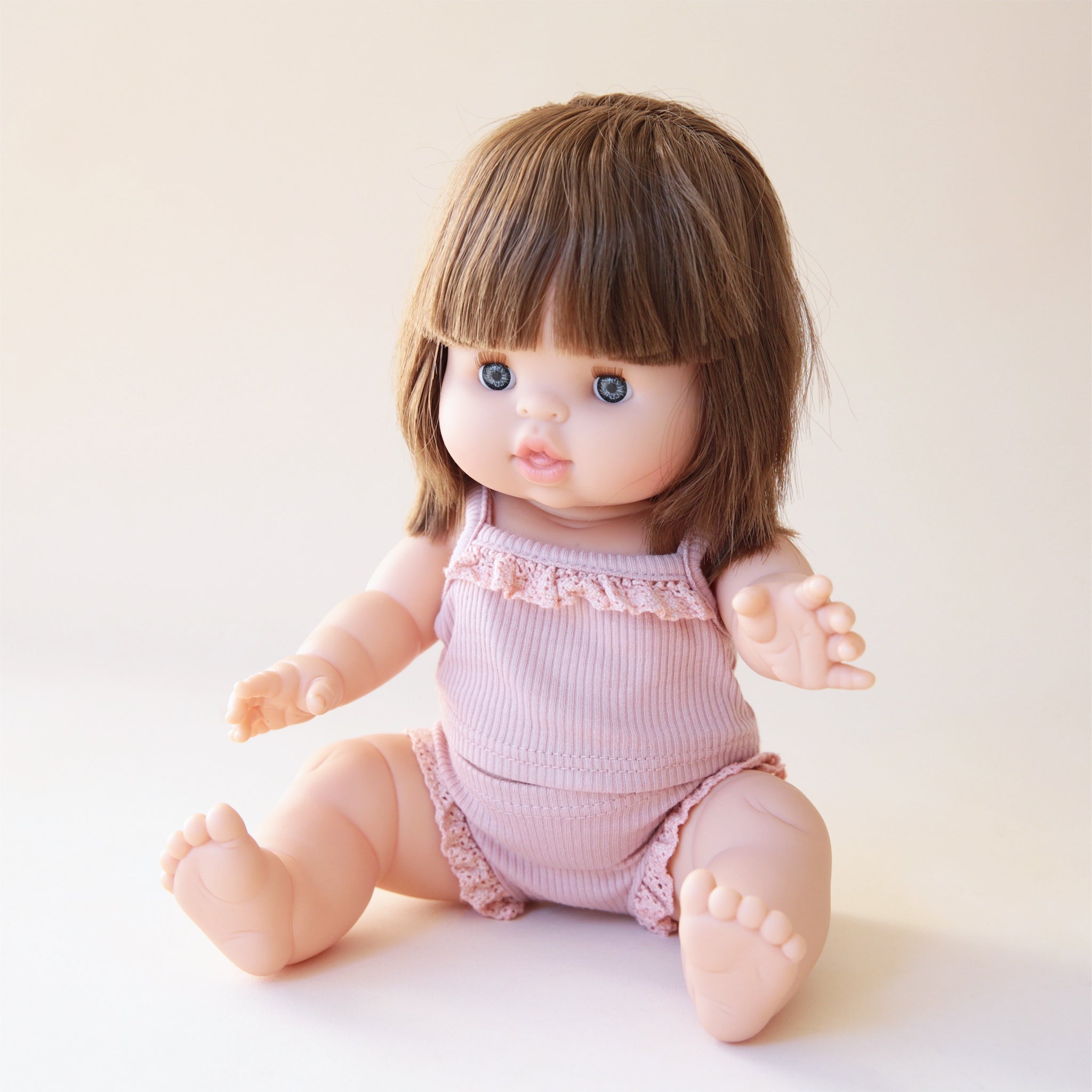 A baby doll with blue eyes, straight dark brown hair that is about shoulder length with bangs and wearing a light pink tank top set not included.