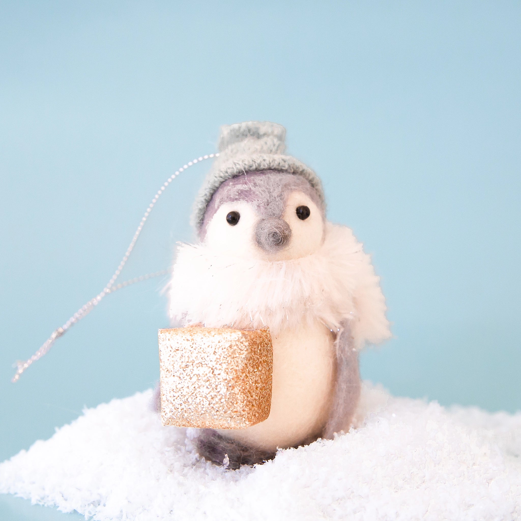 On a blue background is a grey and white felt penguin with a light green beanie and holding a gold gift box in hand. 