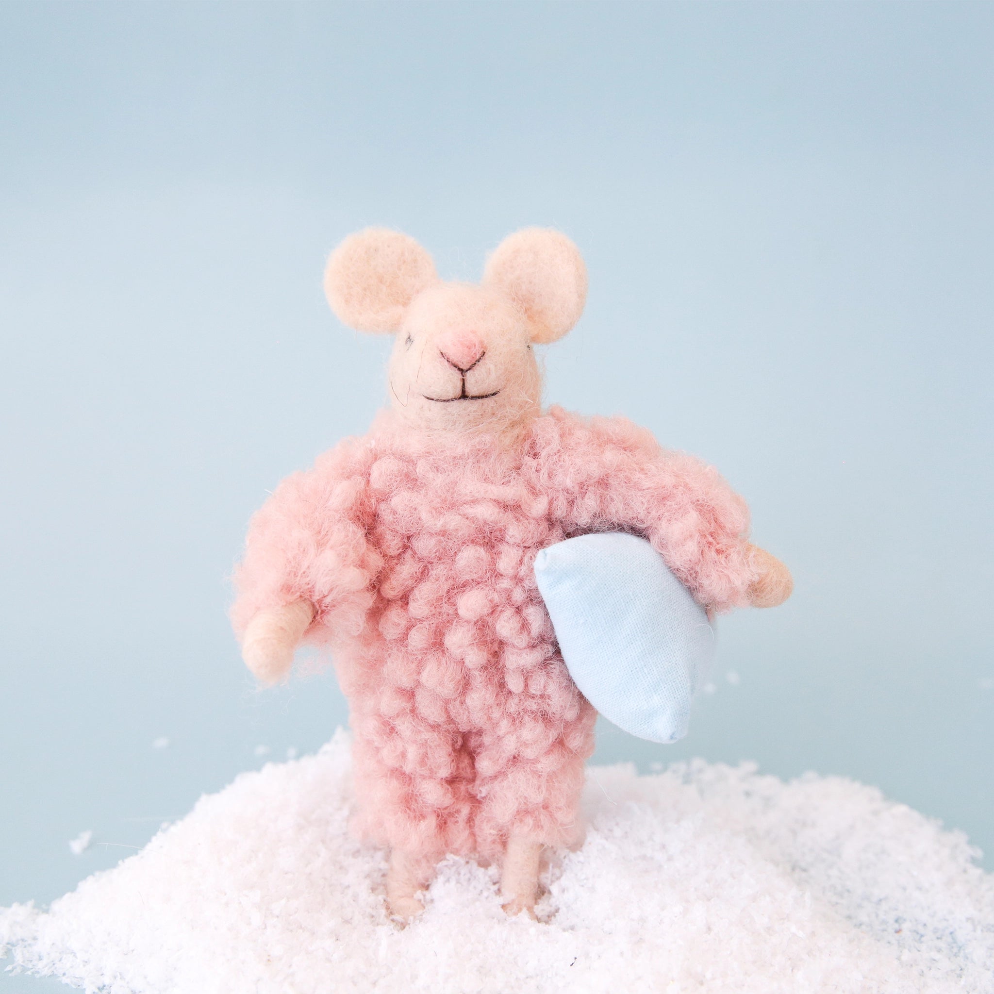 On a blue background is a felt mouse ornament in a cream shade with a fuzzy pink jumpsuit on and holding a pillow. 