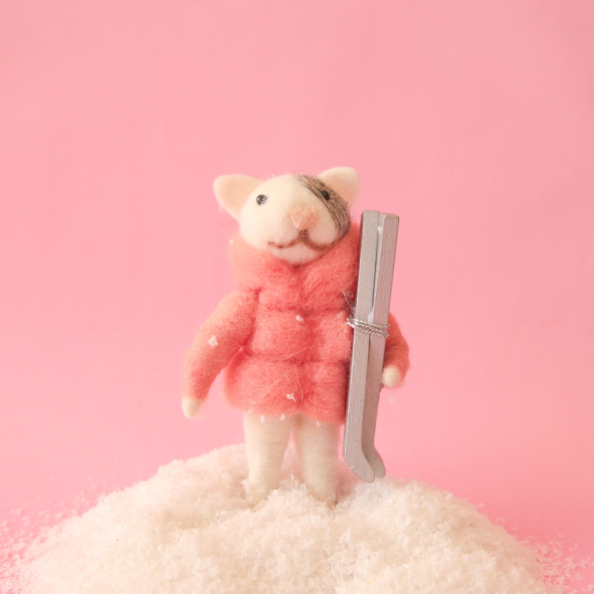 On a pink background is a white and grey felt cat ornament wearing a pink puffy jacket and holding a pair of silver skis