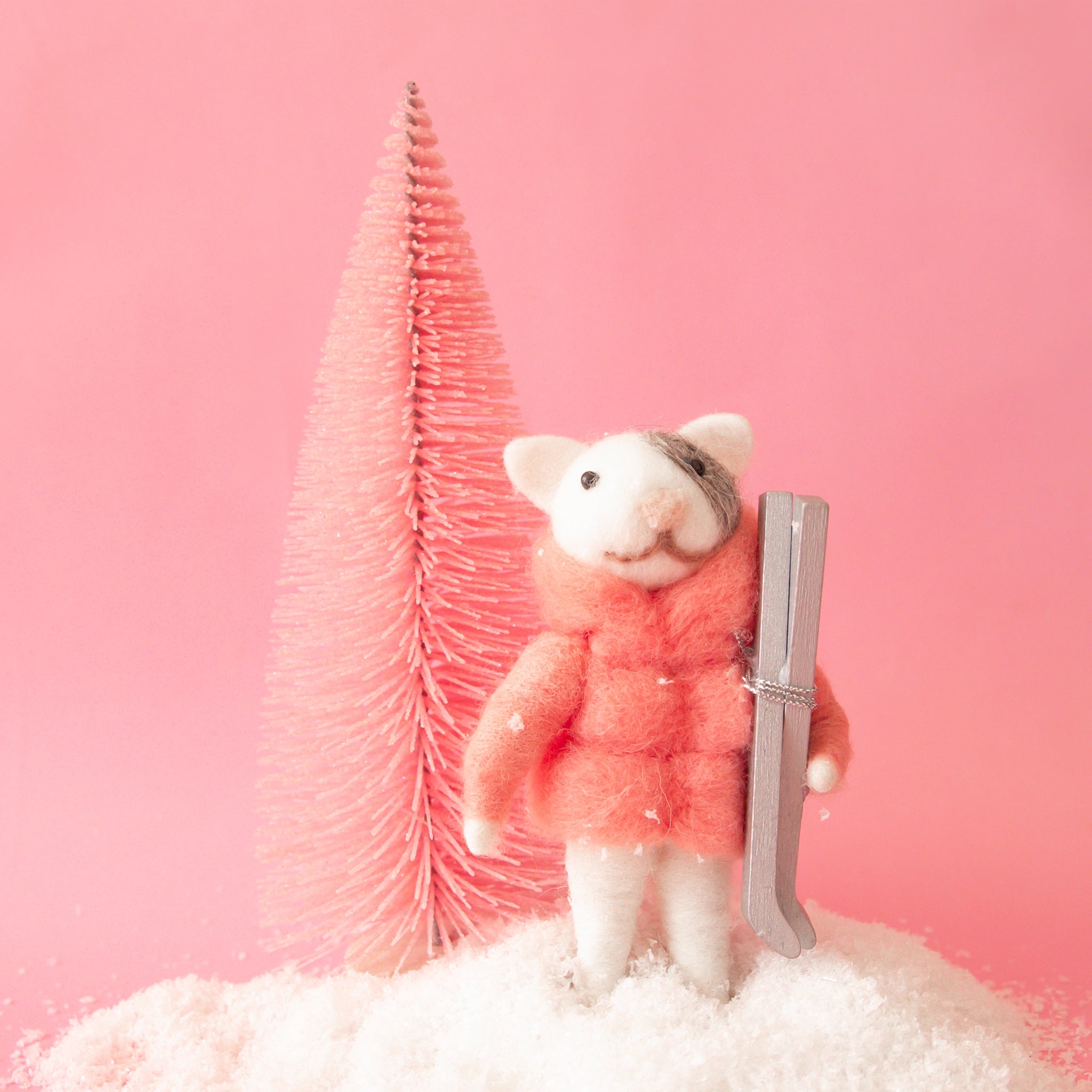 On a pink background with a pink bristle brush tree is a white and grey felt cat ornament wearing a pink puffy jacket and holding a pair of silver skis. 