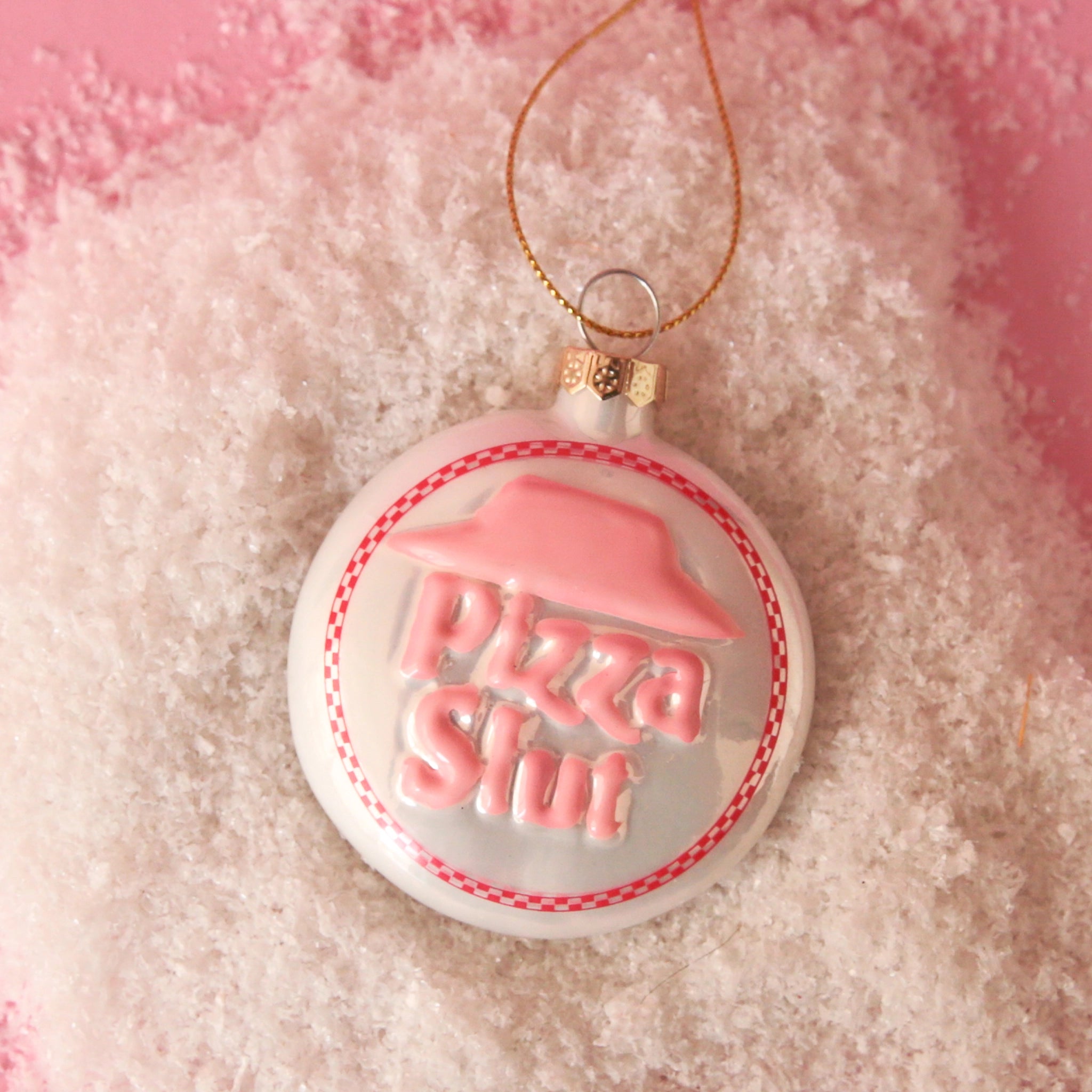 On a pink snowy background is a white and pink circle glass ornament that has text in the center that reads, "Pizza Slut". 