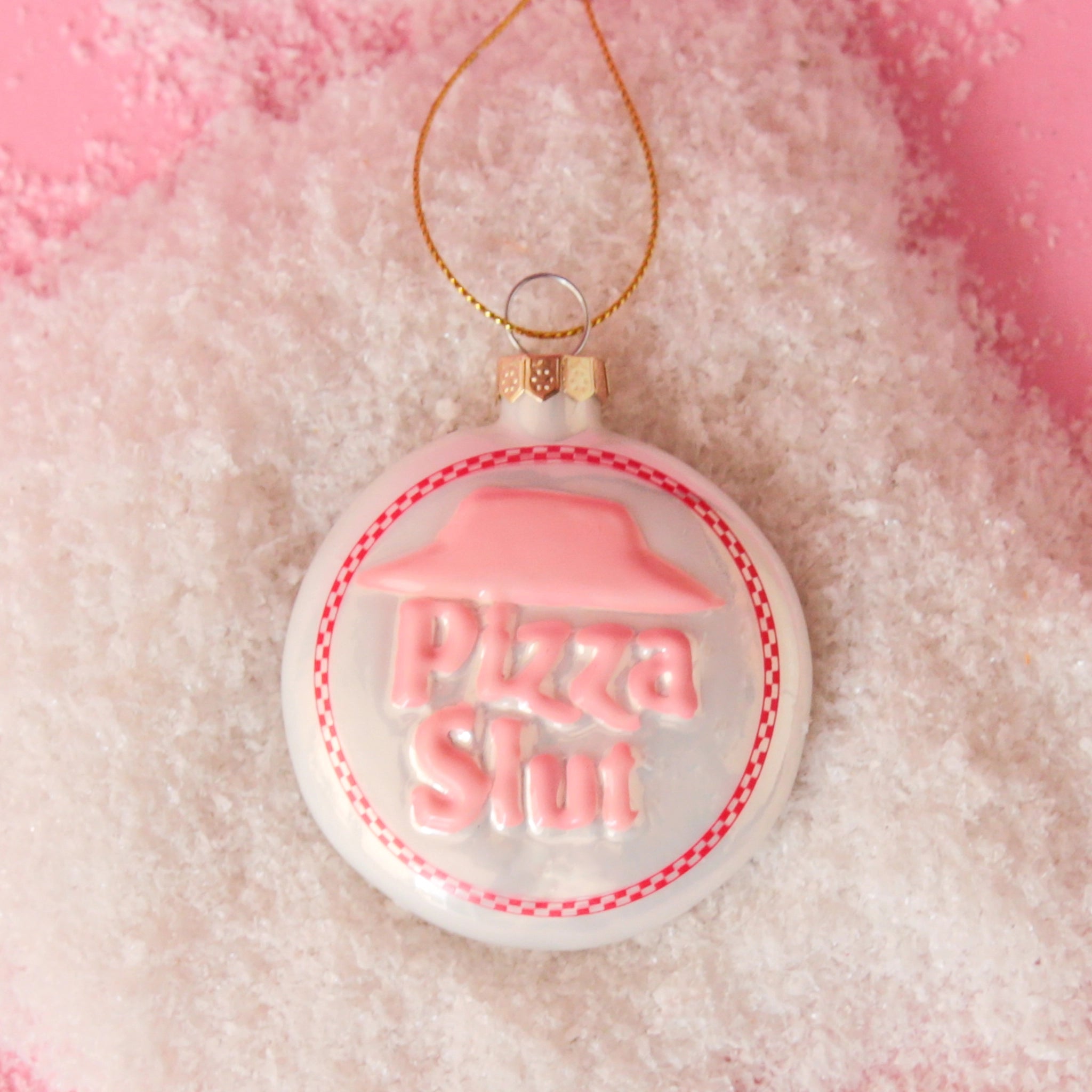 On a pink snowy background is a white and pink circle glass ornament that has text in the center that reads, &quot;Pizza Slut&quot;.