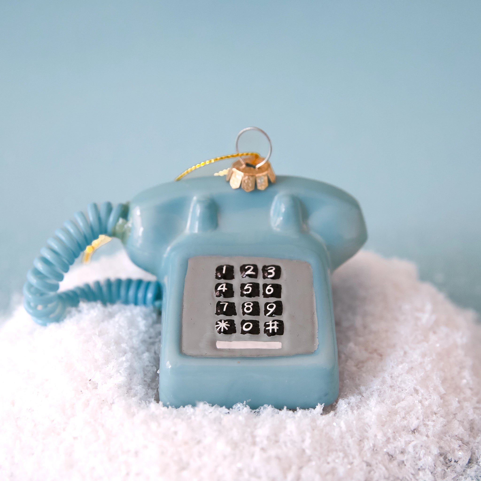 A glass touch tone phone ornament in a shiny blue shade on a snowy blue backdrop.