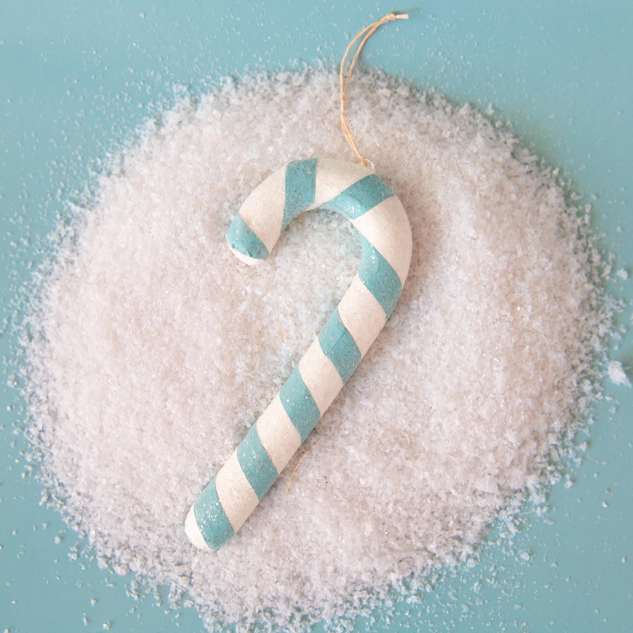 On a blue snowy background is a aqua blue and white striped candy cane ornament. 
