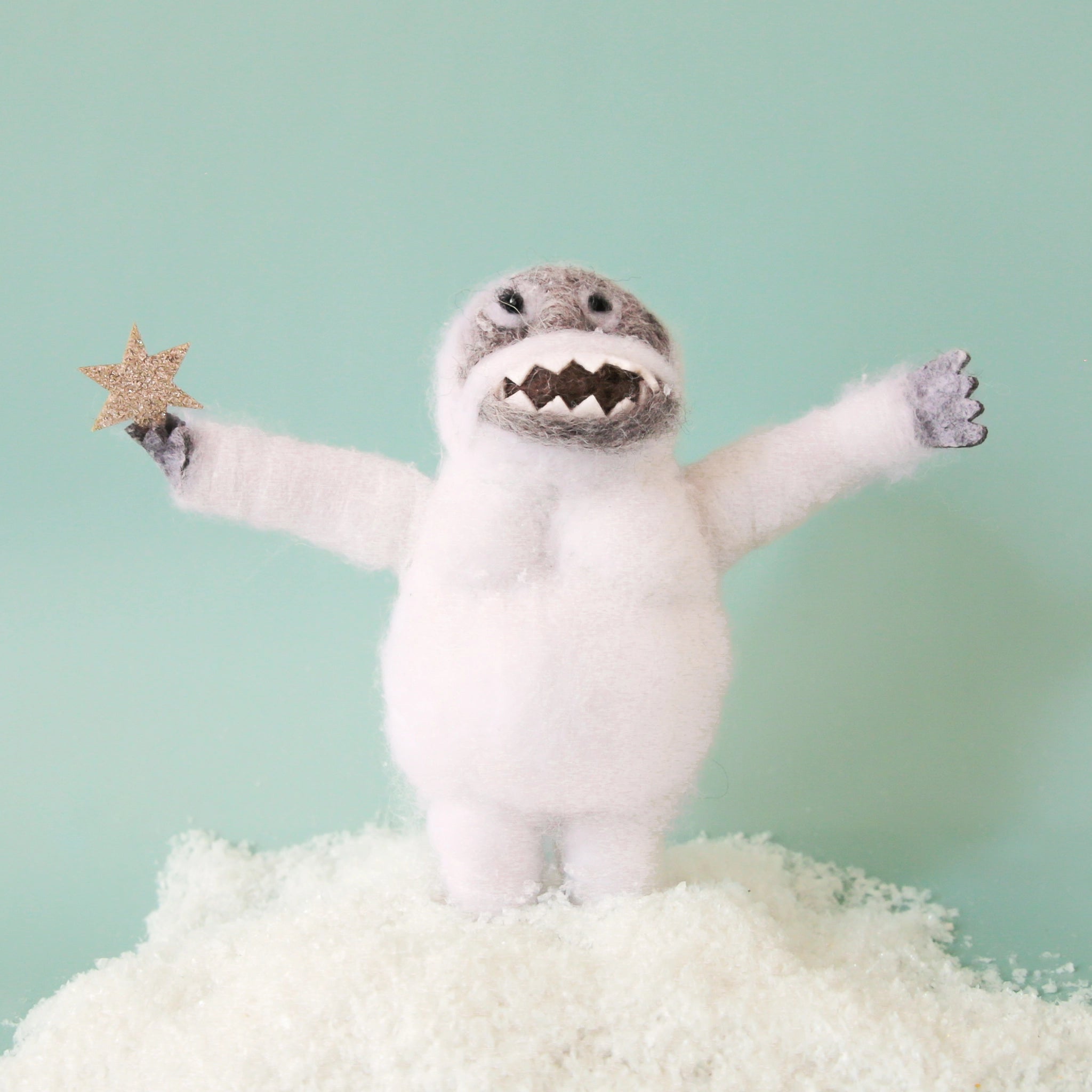 On a light green background is a white and grey felt abominable snowman ornament that&#39;s holding a sparkly star. 