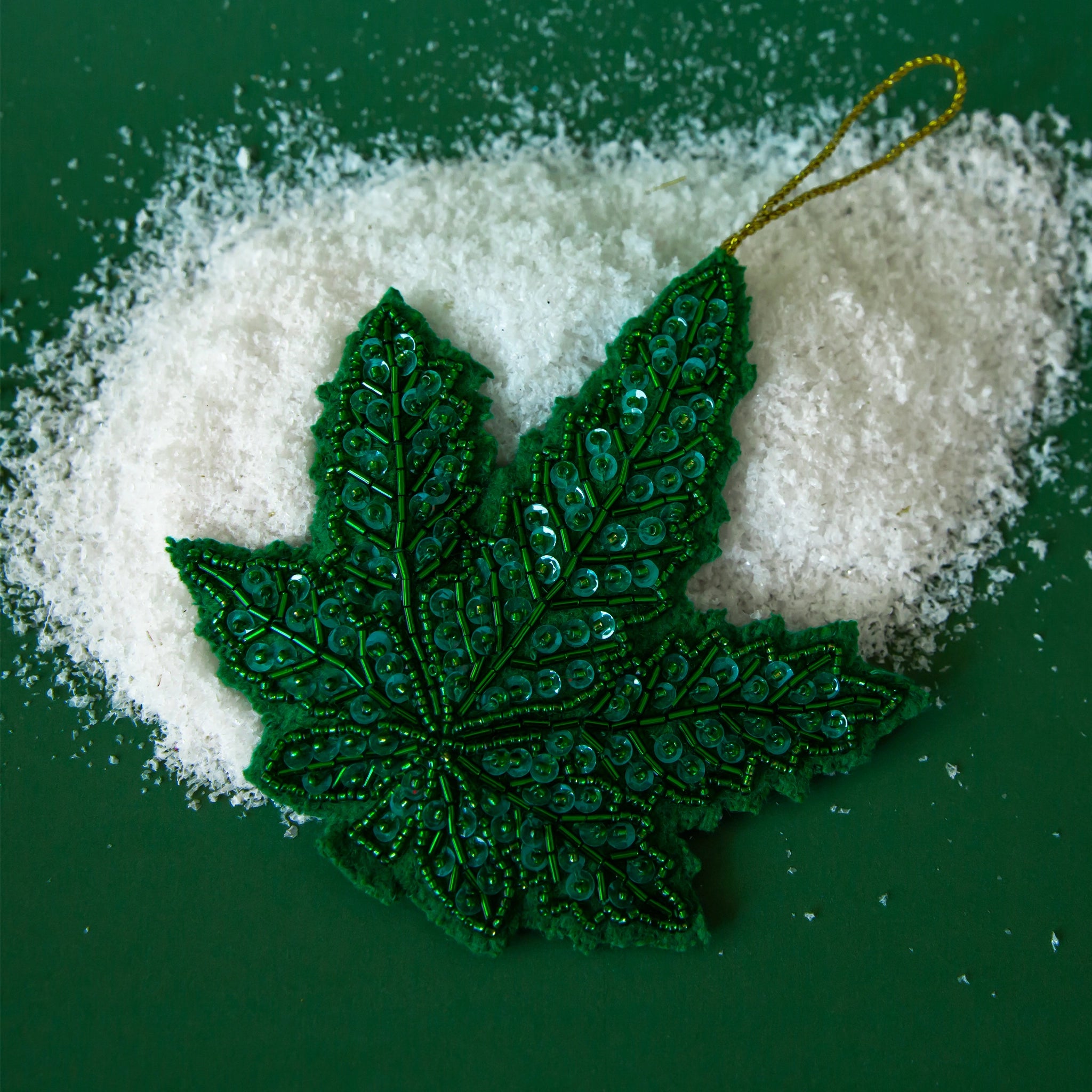 On a green snowy background is a green marijuana leaf ornament with beaded sequin detailing. 