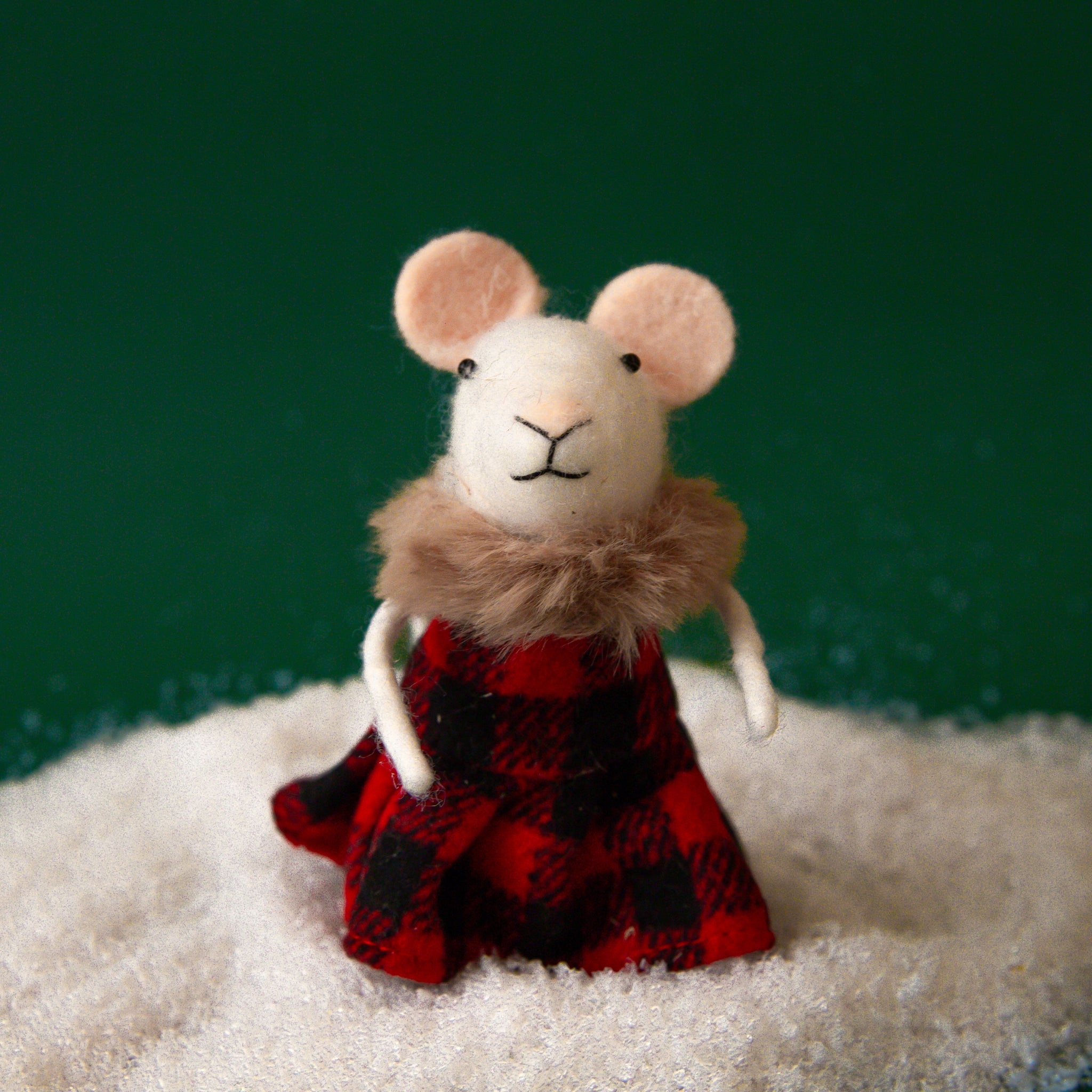 On a green background is a white mouse ornament with a red and black plaid dress on and a brown faux fur collar. 