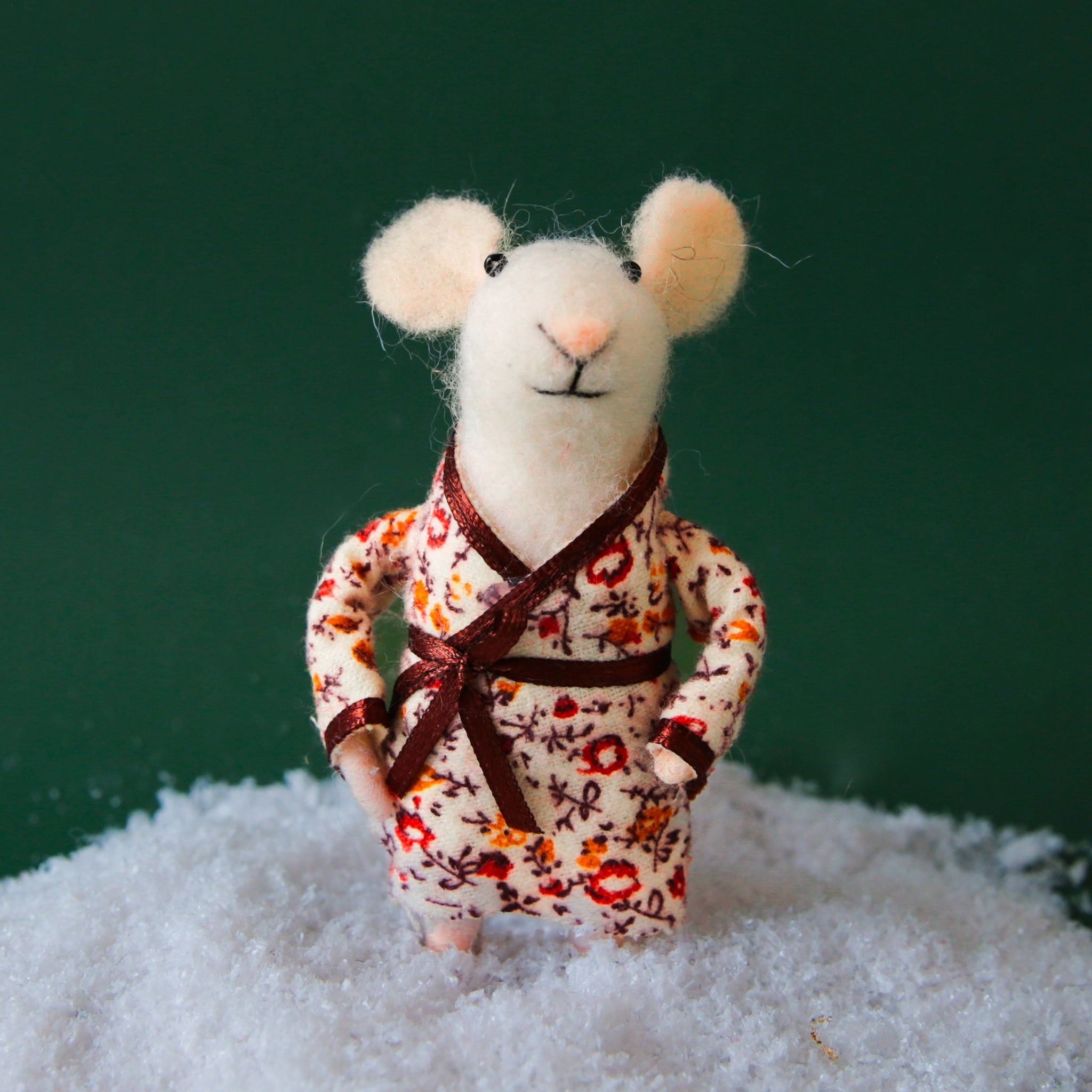 On a dark green background is a white felt mouse ornament with a floral pyjama robe on.