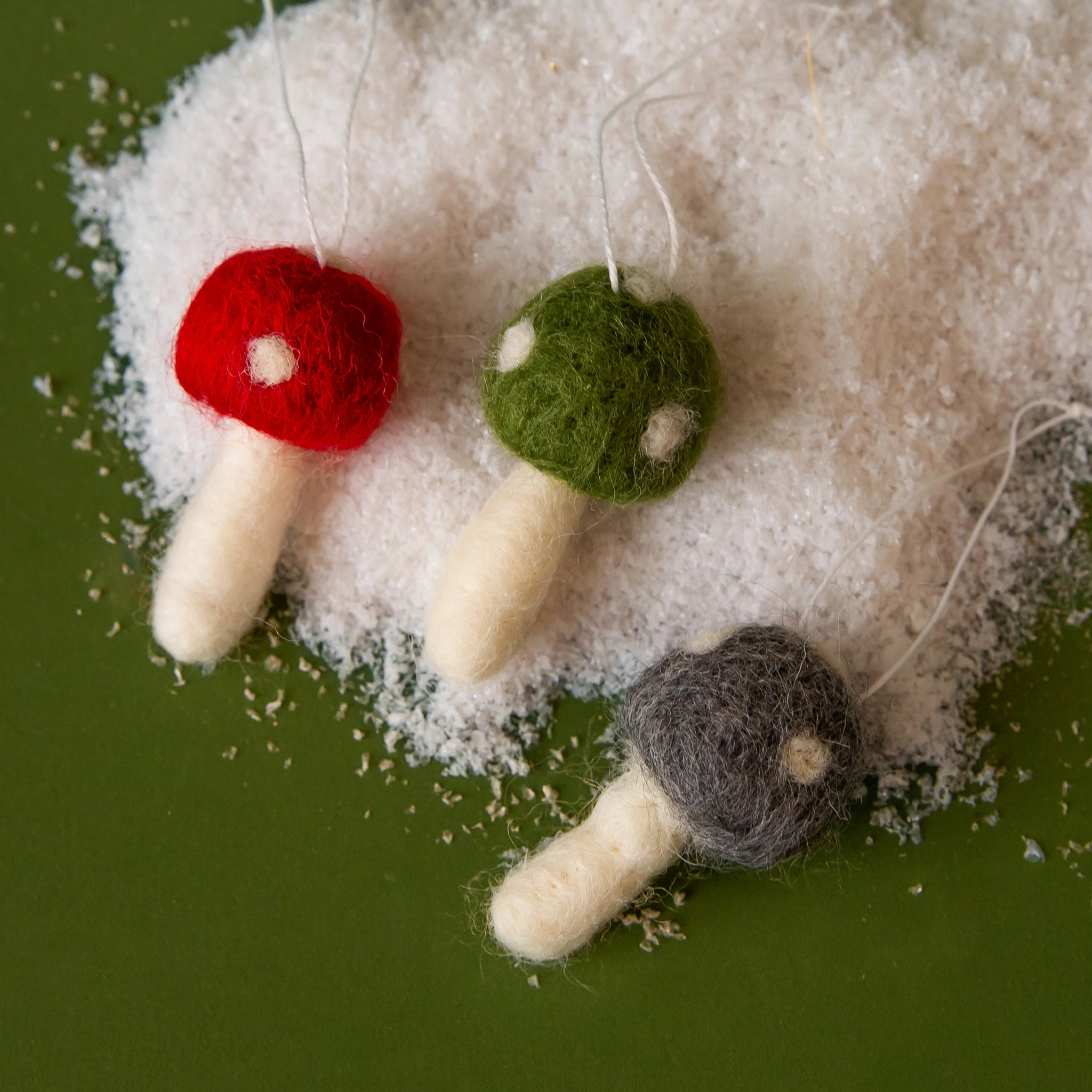 On a green snowy background is three felt mushrooms with a red, green or grey top with white spots. 