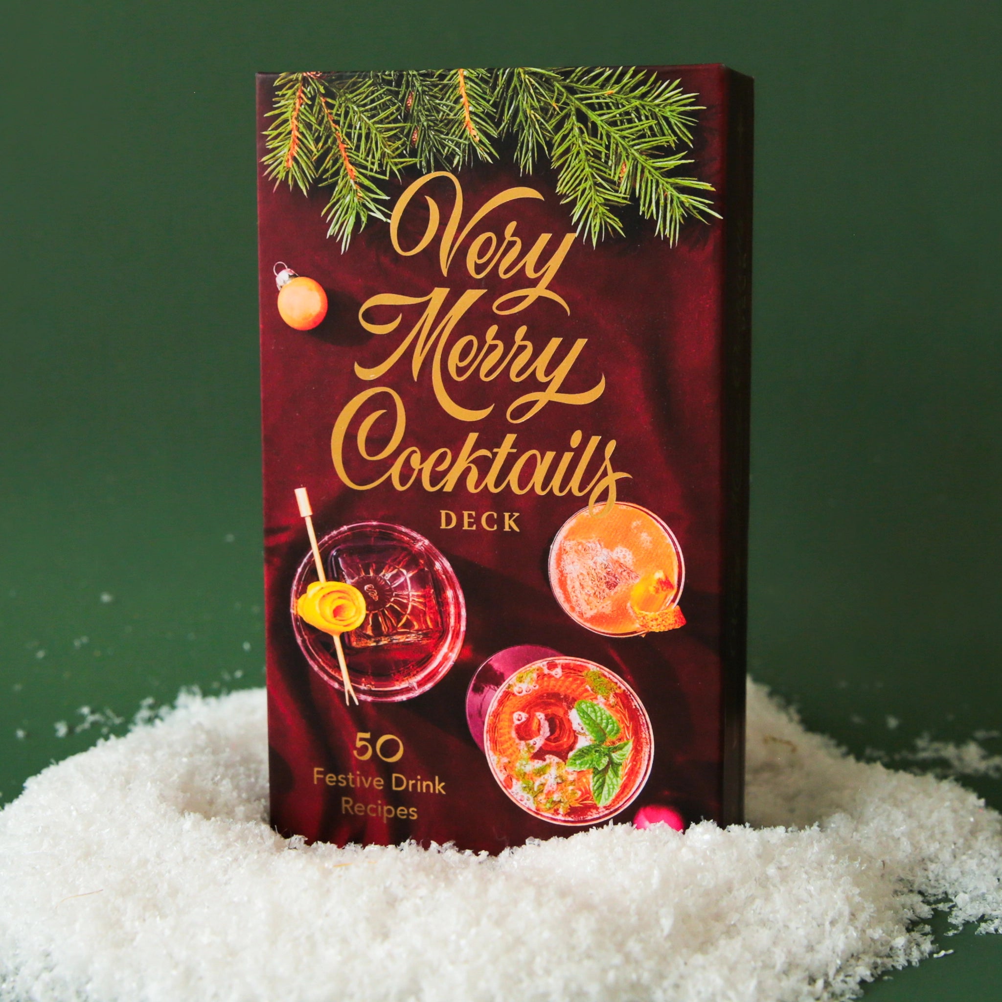 On a green snowy background is a deck of holiday cocktail recipe cards with festive photographs and yellow gold text on the front of the box that reads, "Very Merry Cocktails Deck".