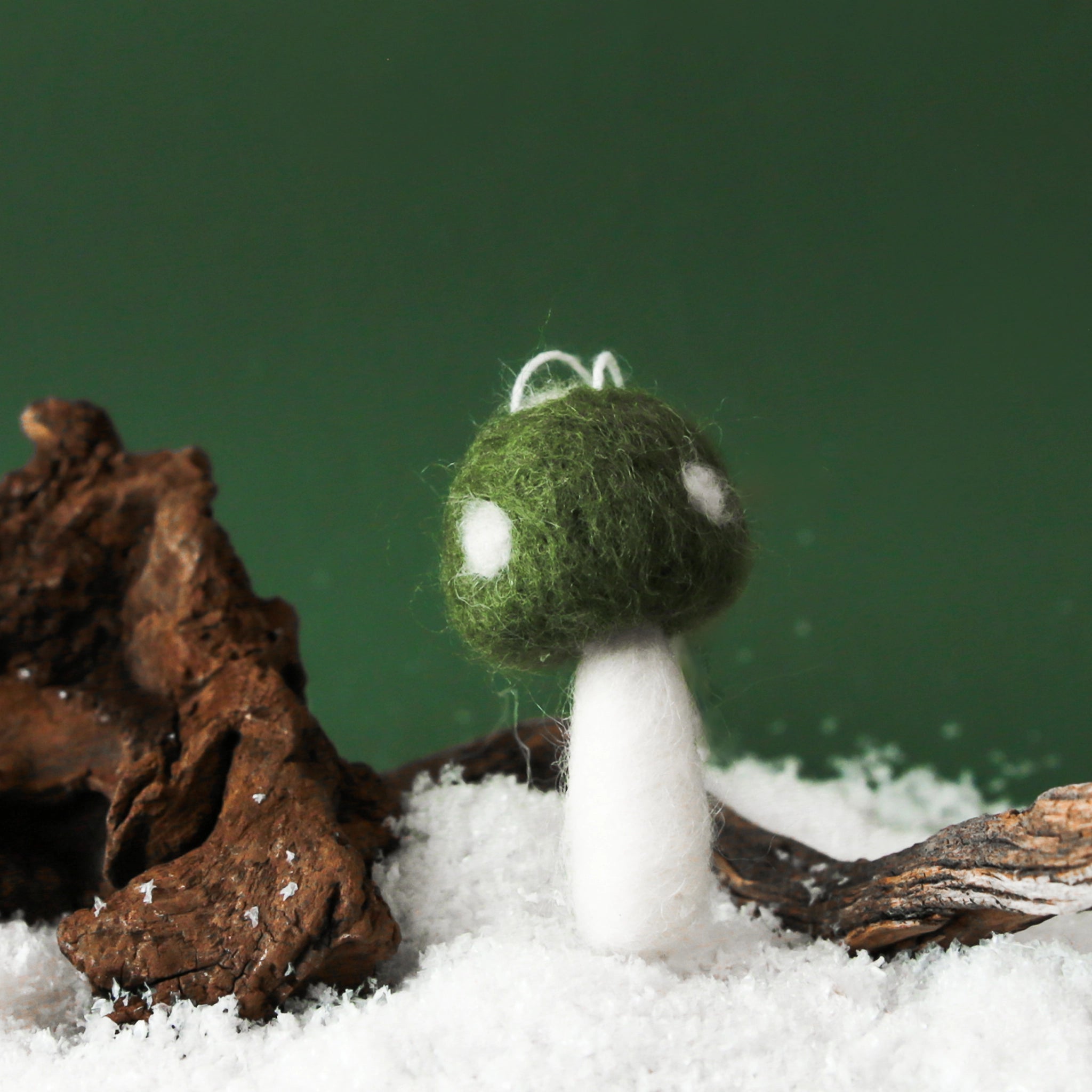 On a green snowy background is a green felt mushroom ornament with ivory spots and details