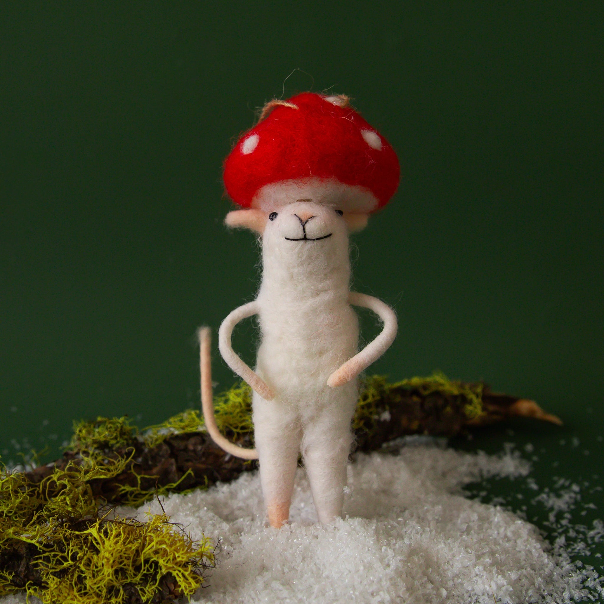 On a dark green background is a white felt mouse ornament with a red mushroom hat on and staged on a snowy and mossy backdrop. 
