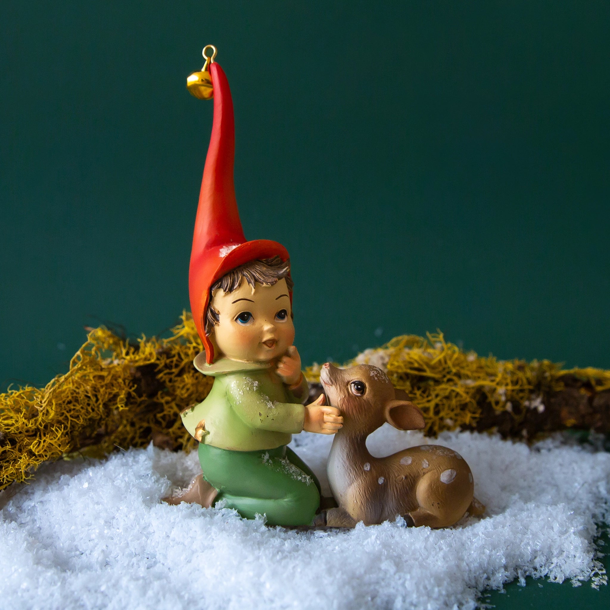 On a green background is an elf figurine with a long red hat and a brown fawn laying next to the elf.