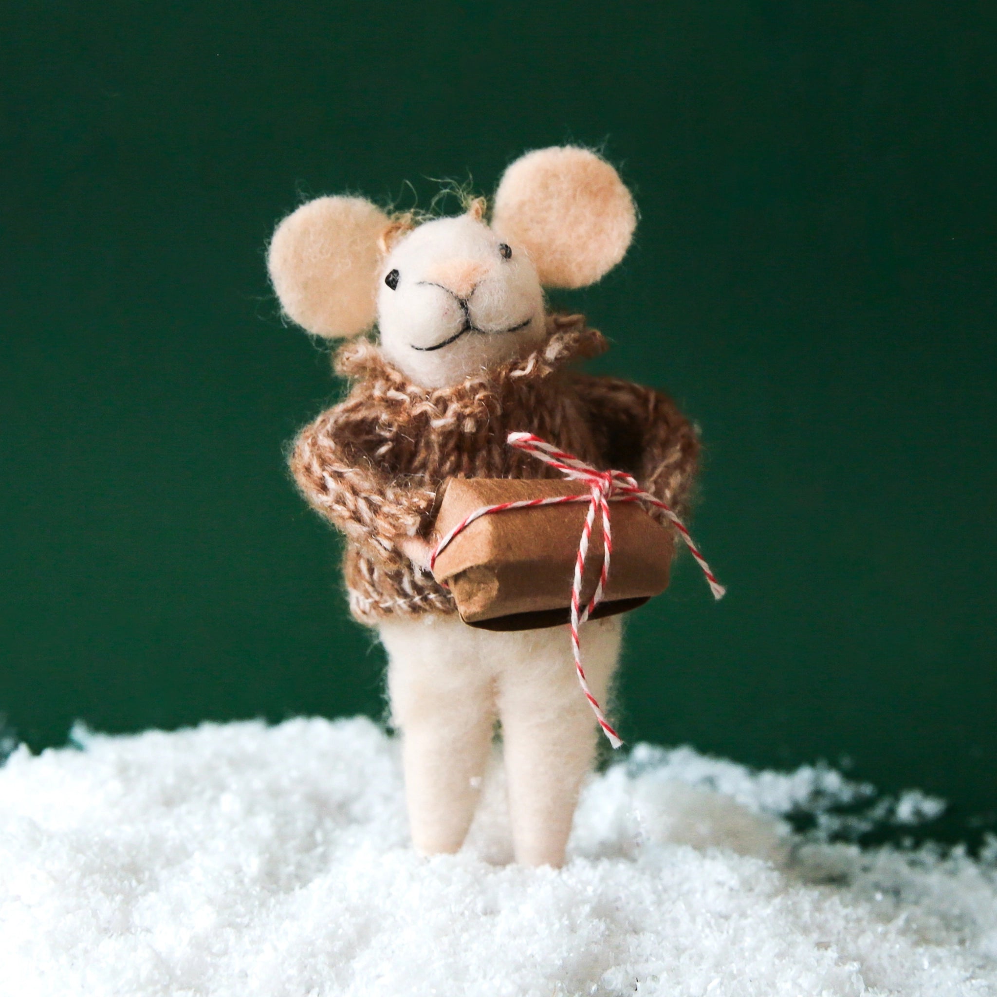 A white mouse ornament with a tan knit sweater and holding a gift wrapped in brown Kraft paper.