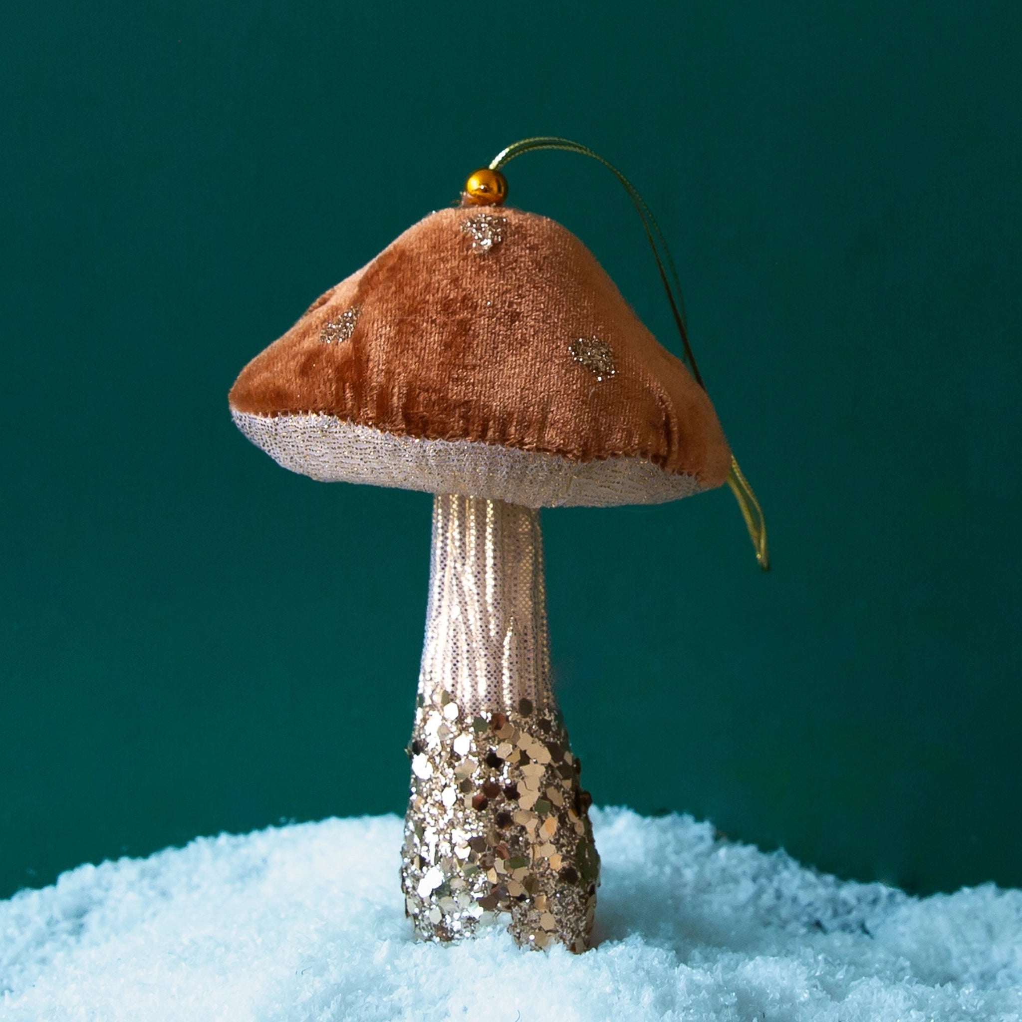 On a green snowy background is a brown and light tan mushroom ornament with gold sparkle details.