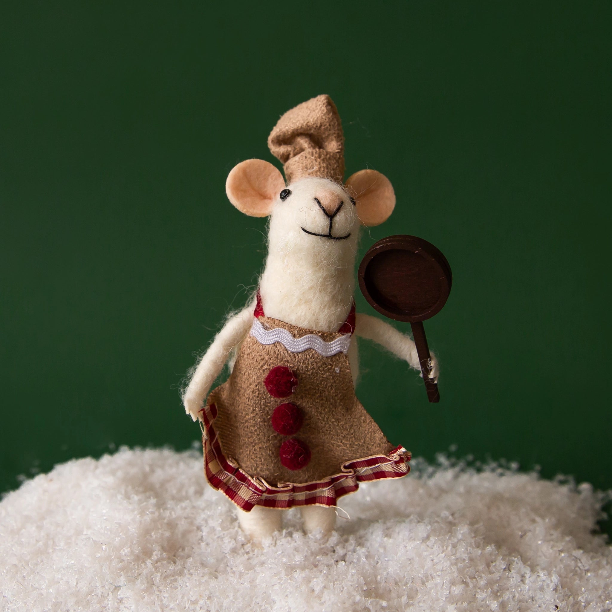On a green background is a white felt mouse ornament with a brown and red apron on with a wood sauce pan in its hand. 