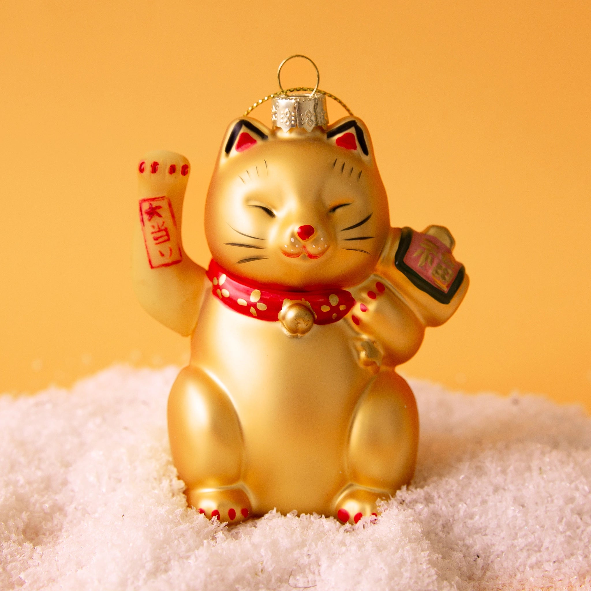 On a yellow background is a gold glass ornament in the shape of a smiling lucky cat.
