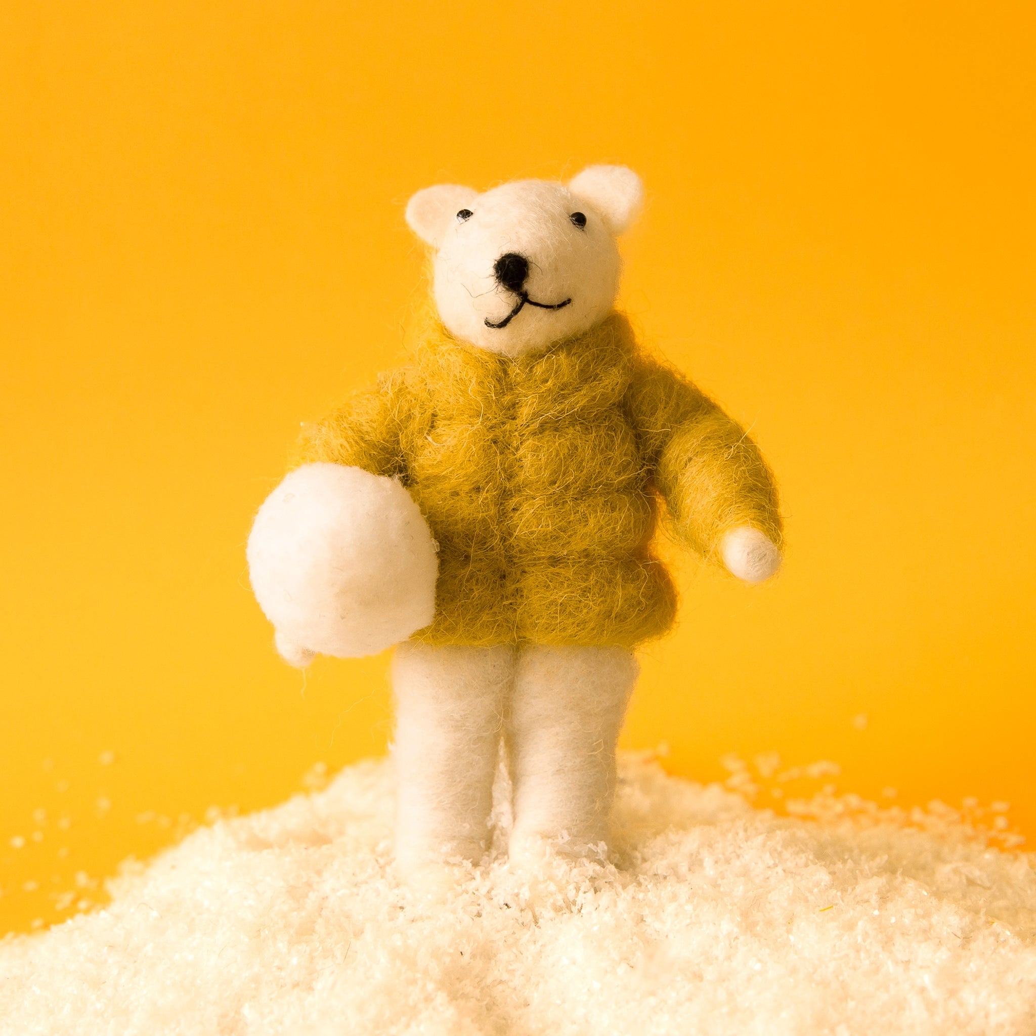 On a yellow background is a white felt bear ornament holding a snowball and wearing a yellow jacket. 