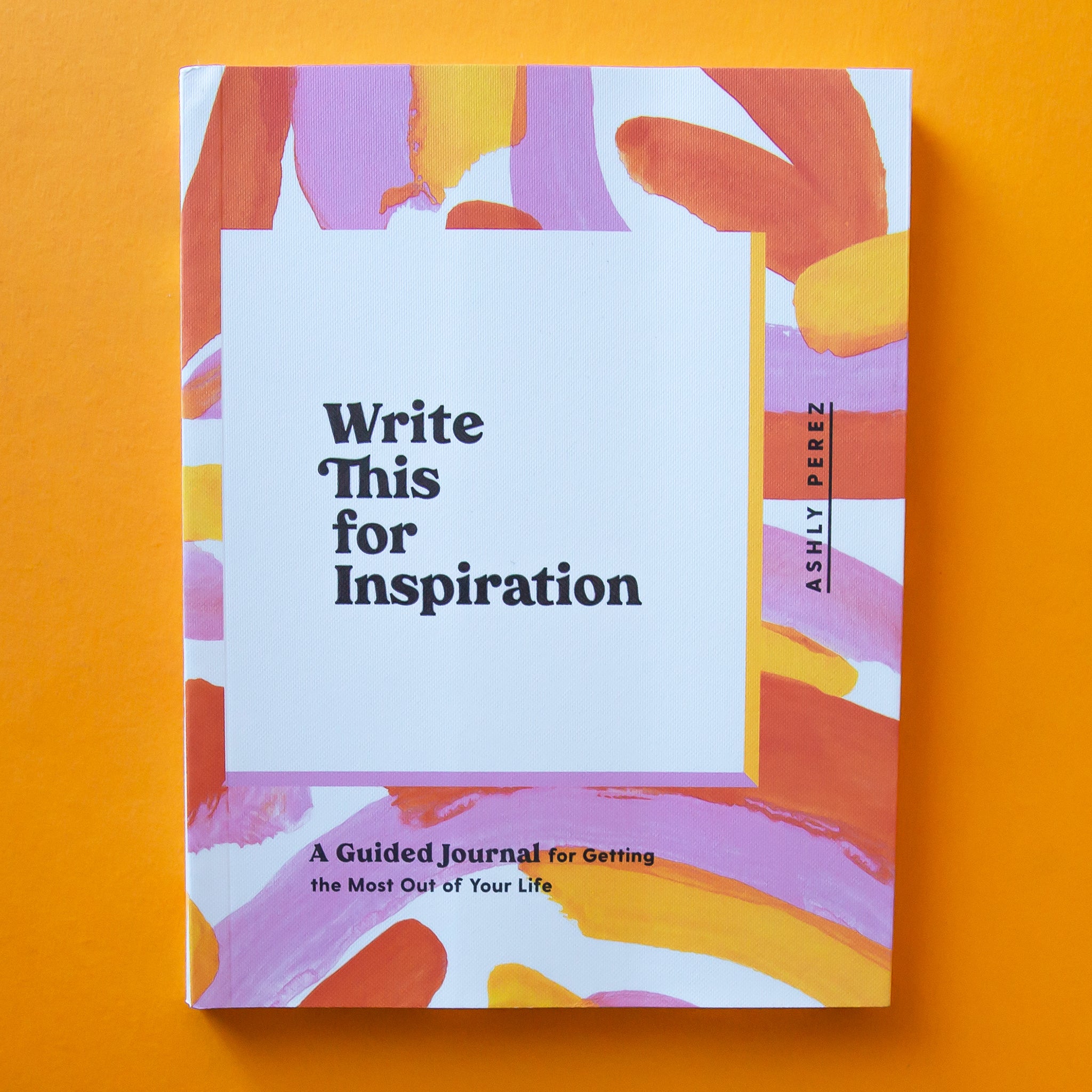 This journal is titled &#39;Write This for Inspiration by Ashly Perez&#39; in black lettering and has an enjoyable free-hand painted design with shades of pinks and oranges on top of a white background. The binding of the book is also white with the title spelt out in similar colorful lettering.