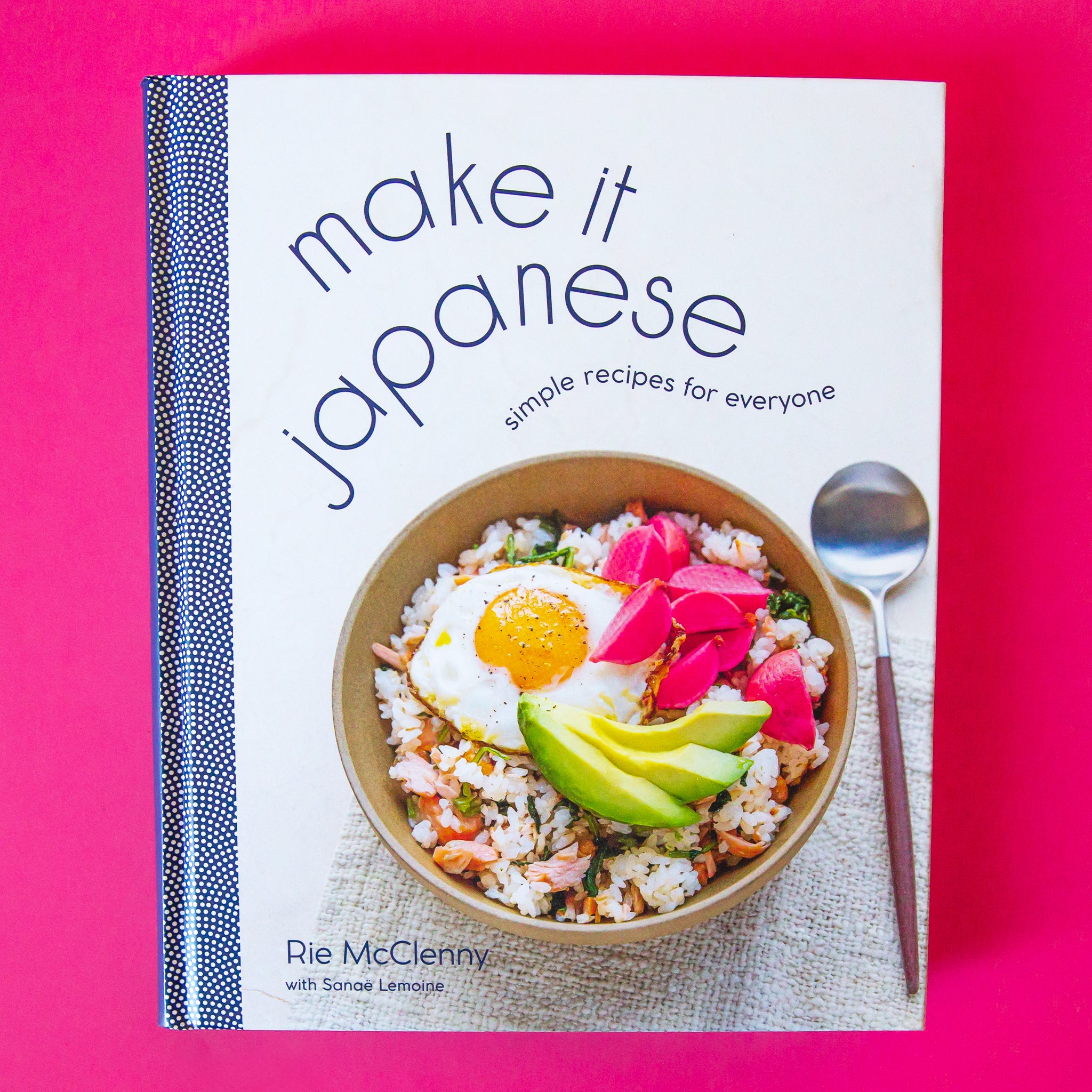 On a pink background is an ivory book cover with a book of food and wavy text above it that reads, &quot;make it japanese simple recipes for everyone&quot;.