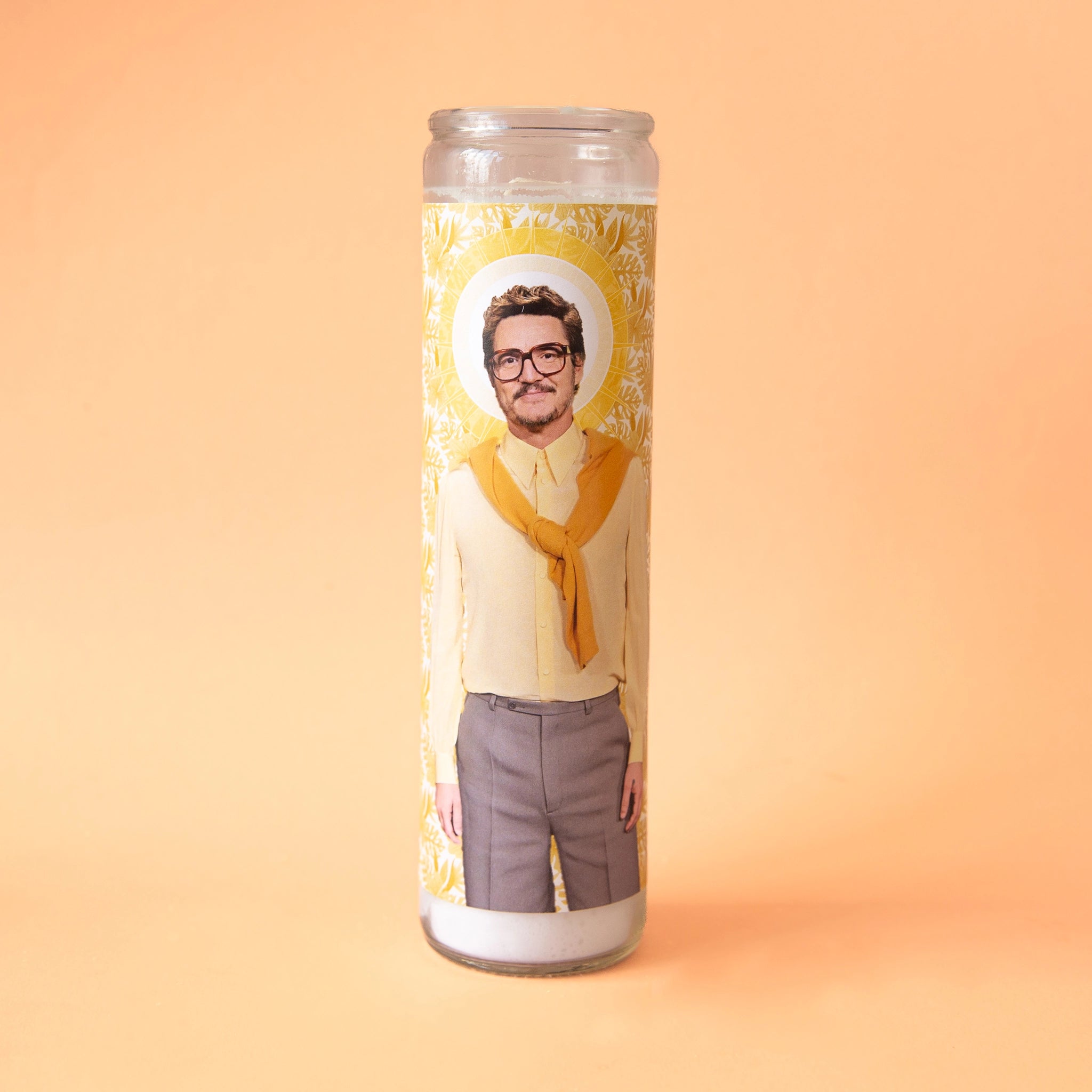 On a yellow background is a tall thin prayer candle in a clear glass jar with a label on the front featuring a photograph of Pedro Pascal in glasses and wearing grey slacks a yellow button down and a orange sweater tied around his shoulders.