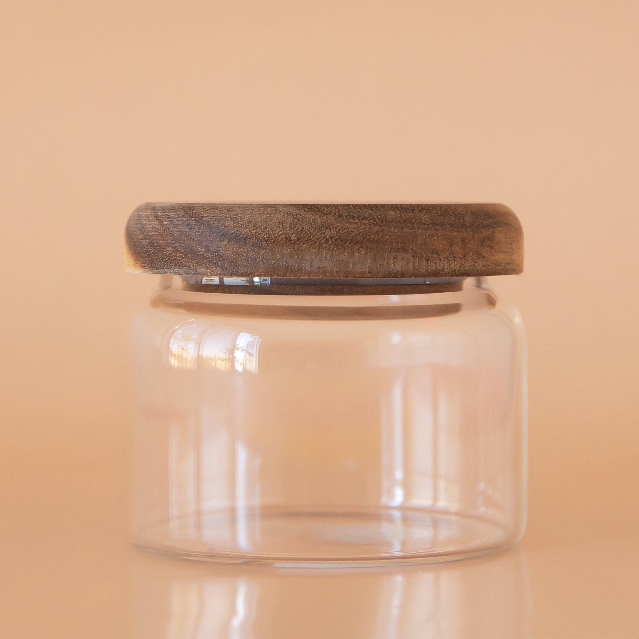 On a peach background is a clear glass storage canister with an acacia wood lid.