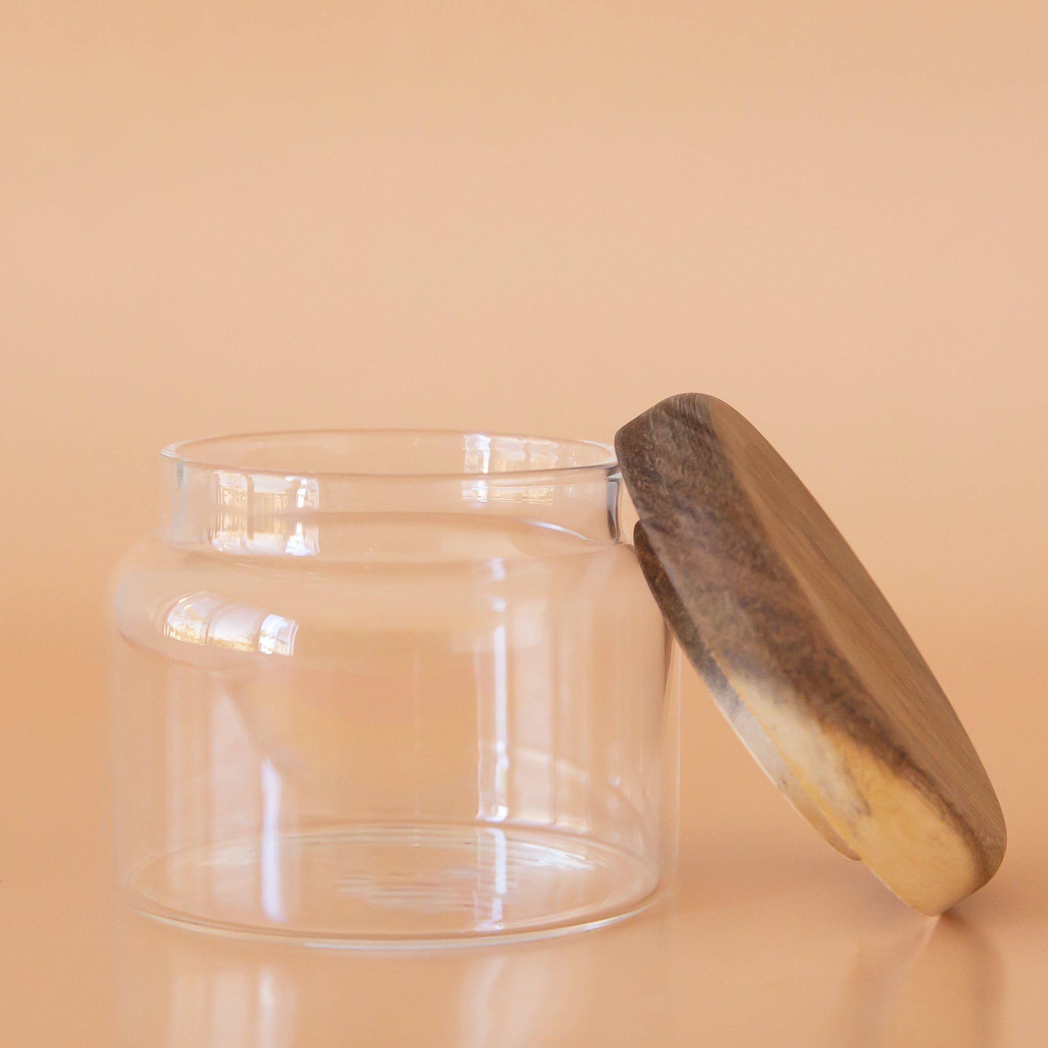 On a peach background is a clear glass storage canister with an acacia wood lid.