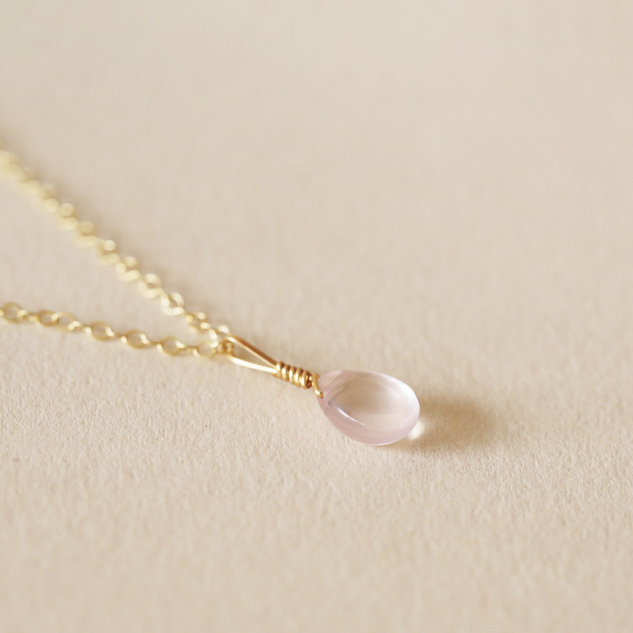 On a neutral background is a gold chain necklace with a rose quartz stone in the center. 