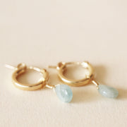 On a neutral background is a gold pair of huggie hoop earrings with a blue Aquamarine dangle deal. 